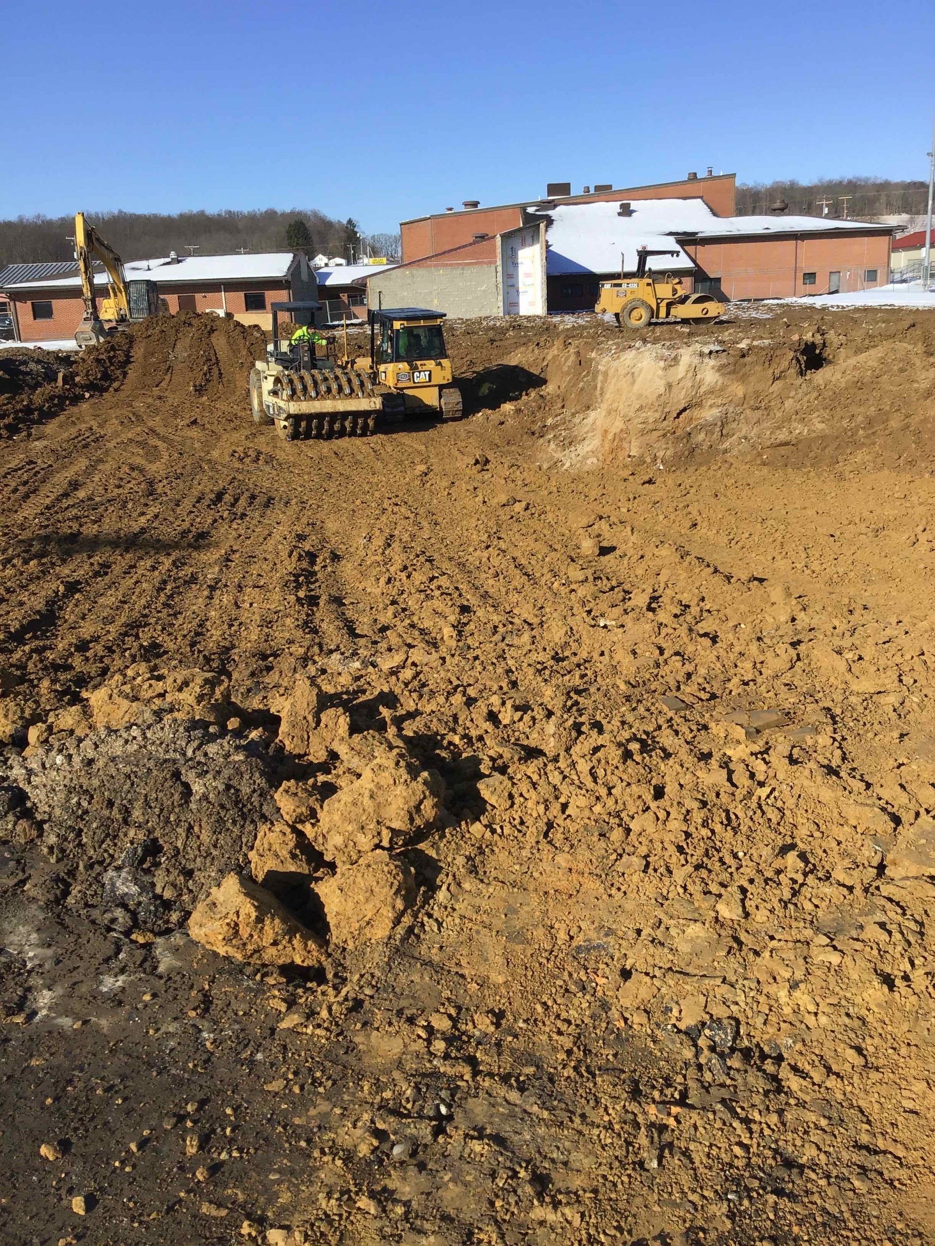 Construction site with earthmoving equipment and brown dirt. Buildings in the background. Sunny day.