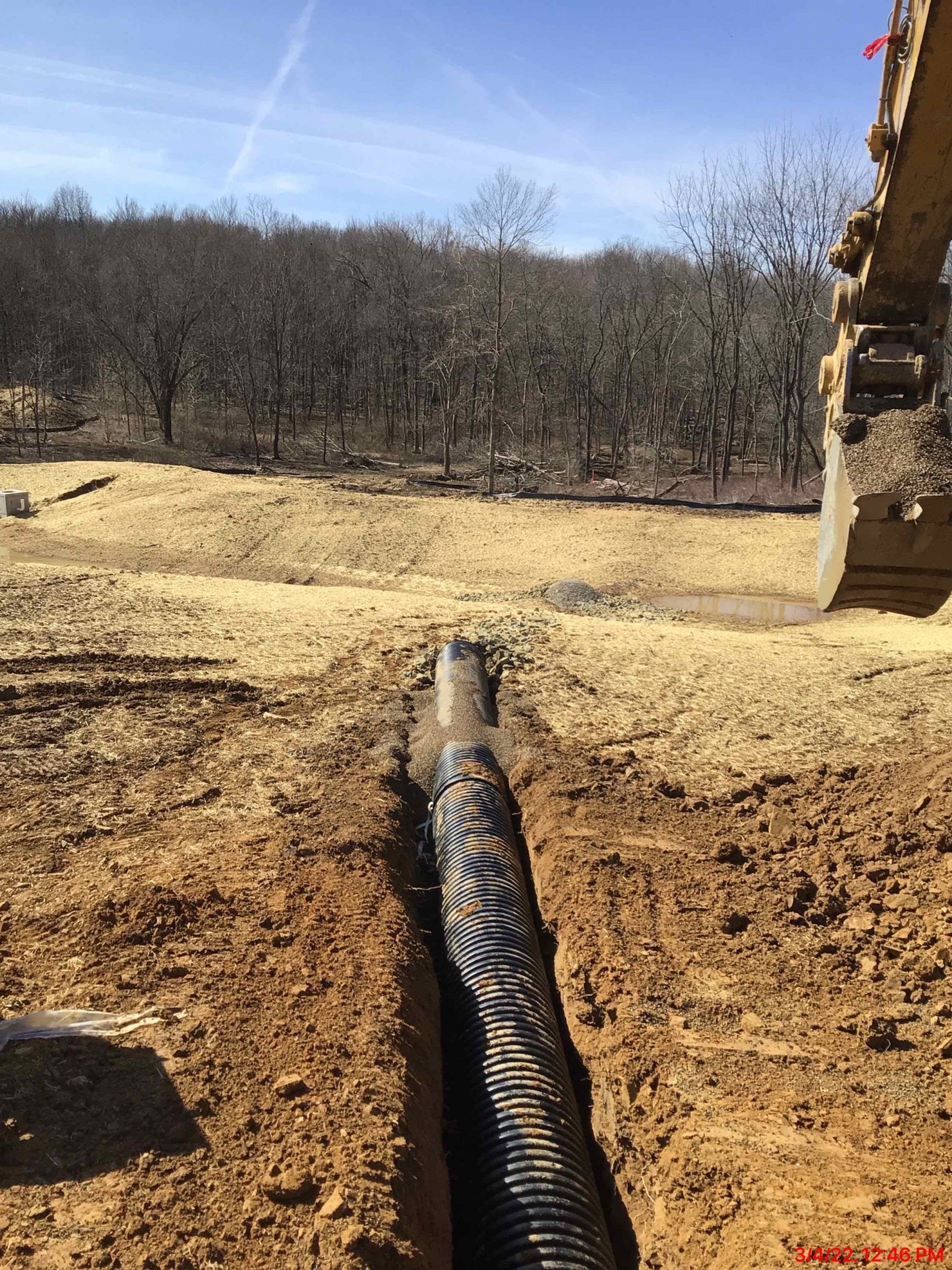 Black corrugated pipe in a trench on a construction site. Excavator visible to the right, trees in the background.