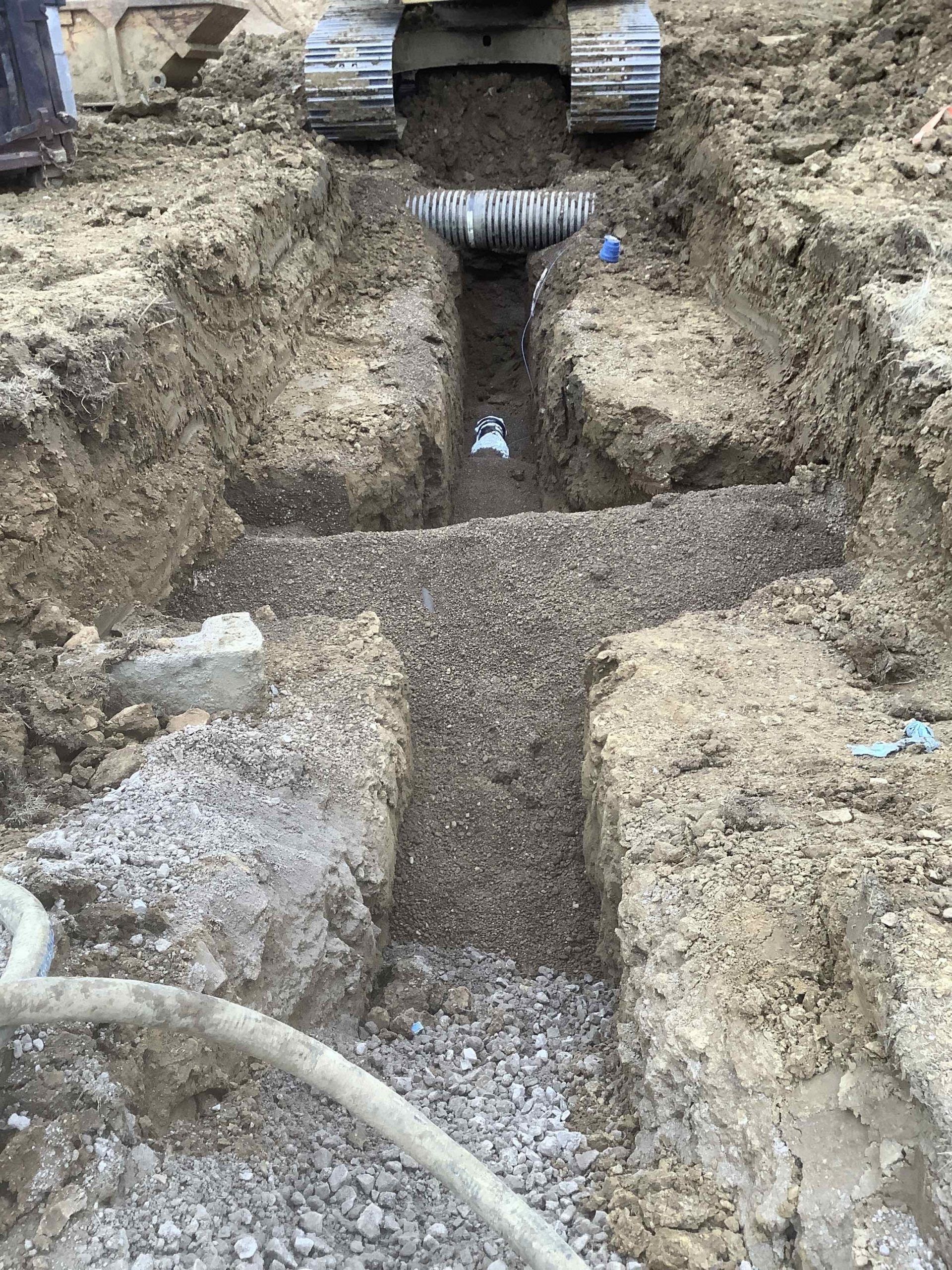 Excavator above a trench; gravel bed; corrugated pipe; dirt and rocks.