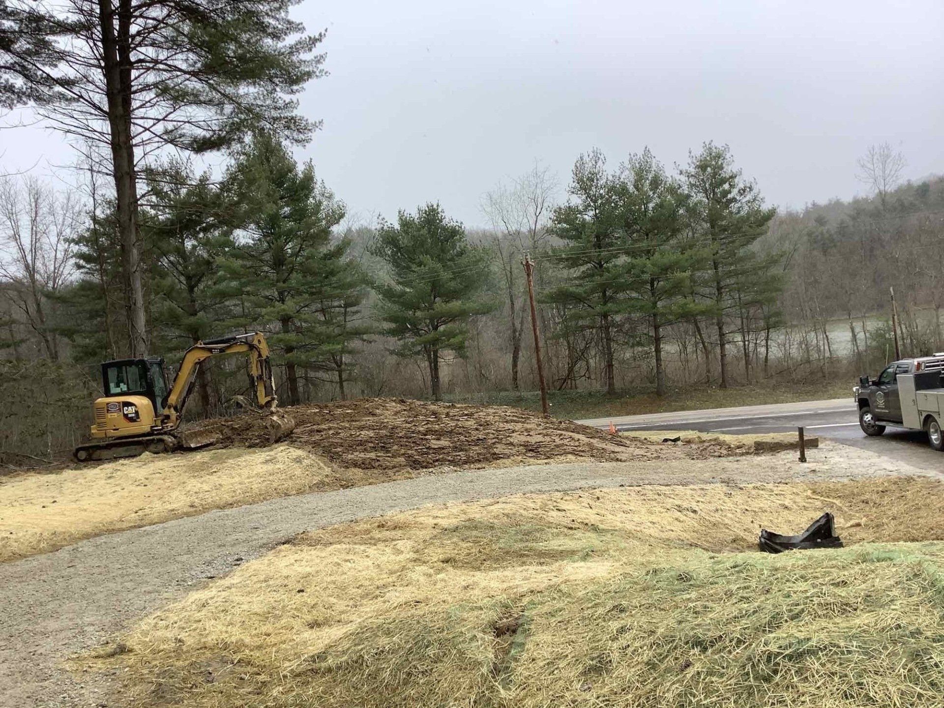 Yellow excavator moving wood chips next to gravel path and road. Trees and vehicle in background.