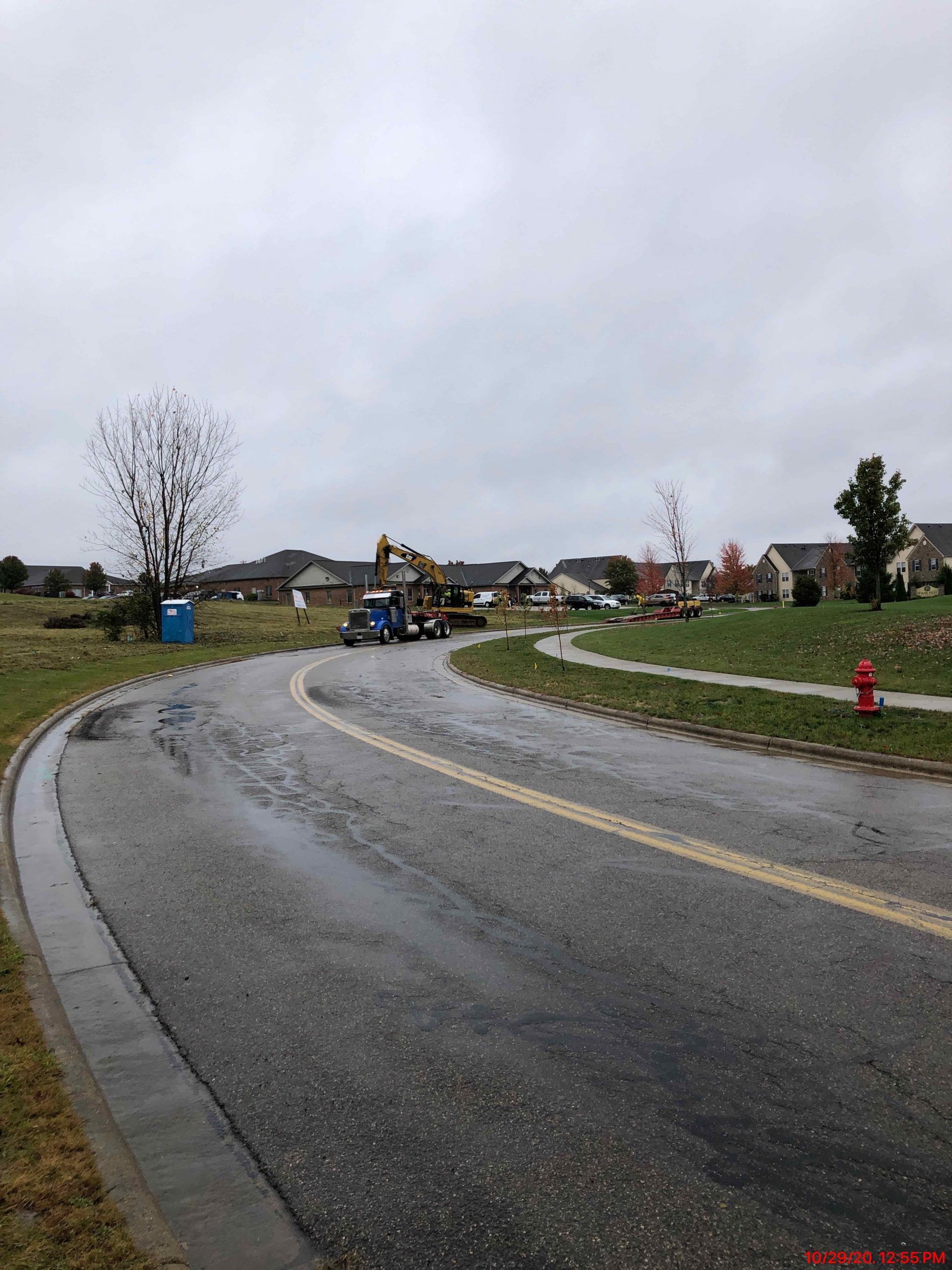 Road construction with equipment, a porta-potty, and houses on a cloudy day.