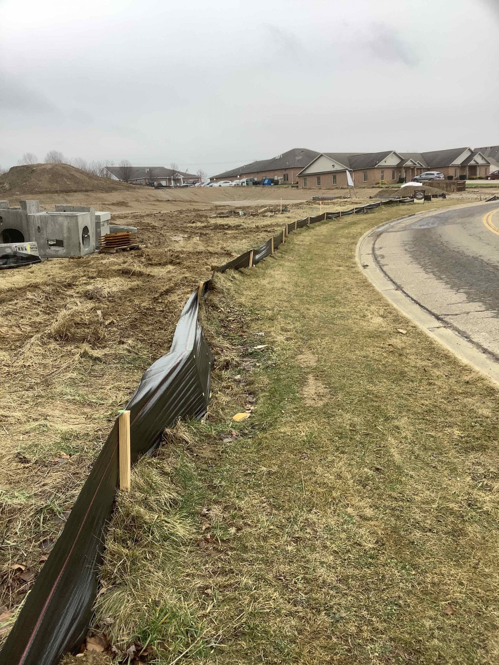 Construction site with a black barrier along a curved grassy area next to a road, and homes in the distance.