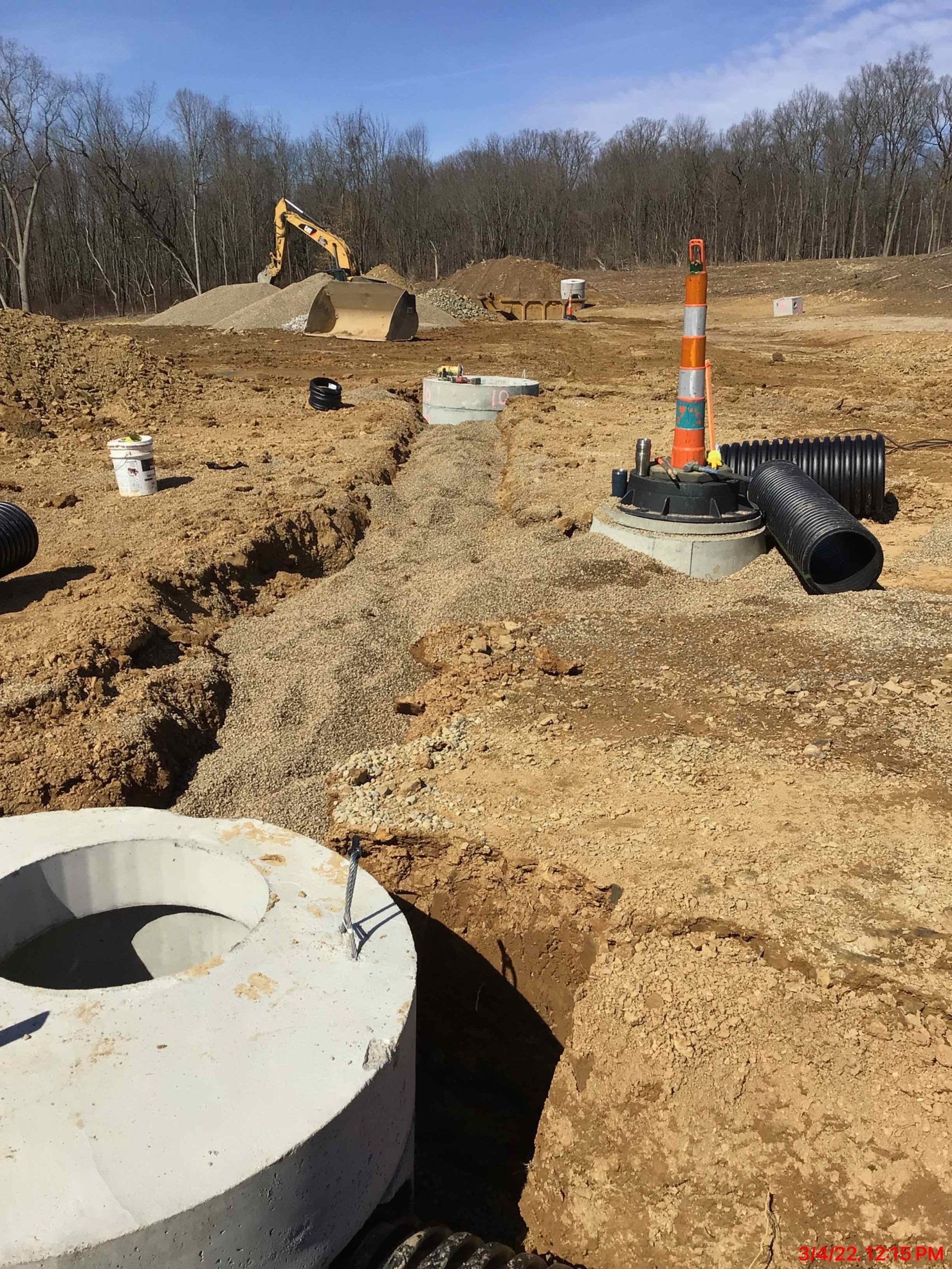 Construction site: excavation with concrete rings, pipes, and an excavator in the background. Sunny day.