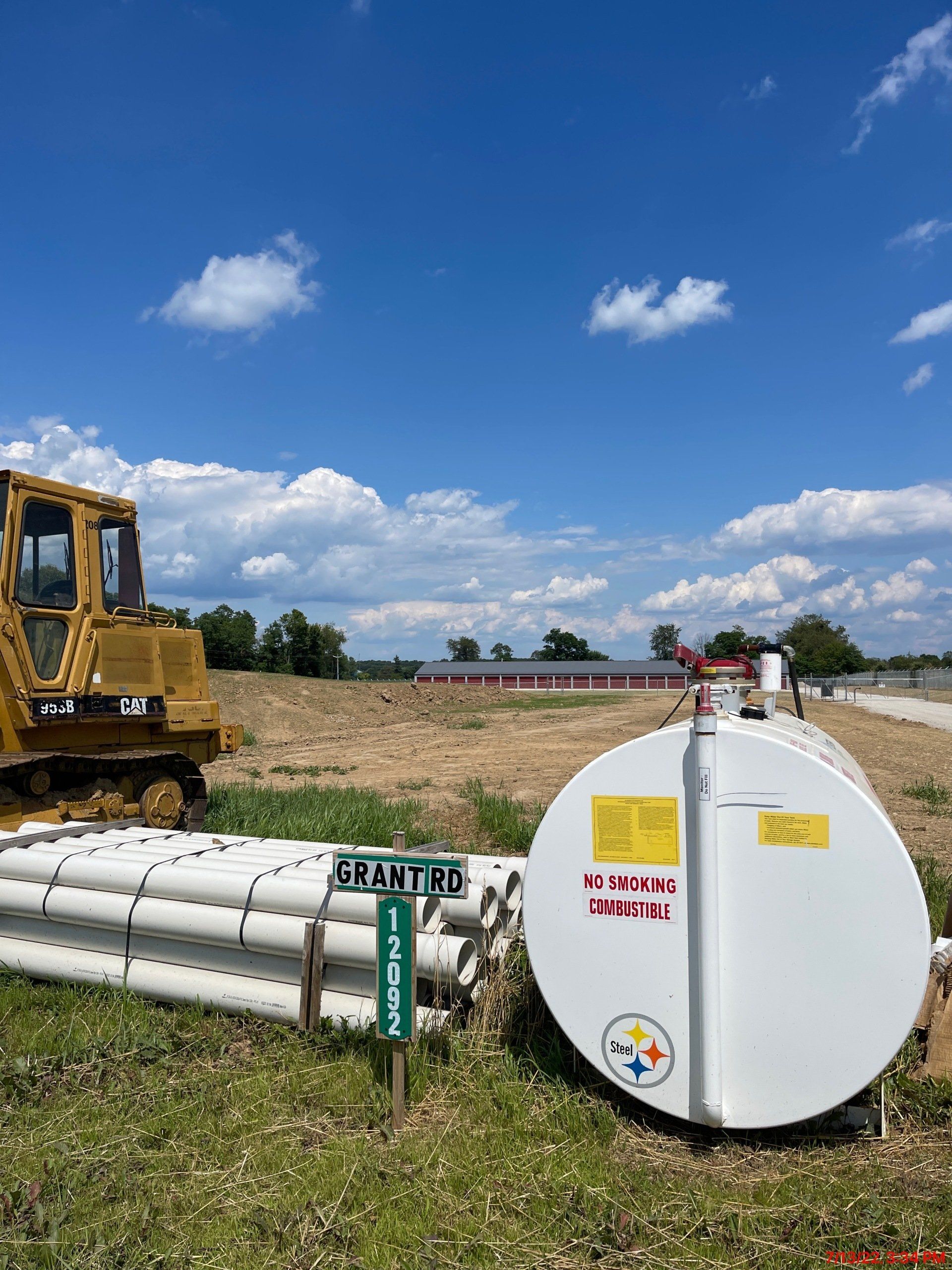 Bulldozer and white tank on a field with stacked pipes and a sign, under a blue sky.