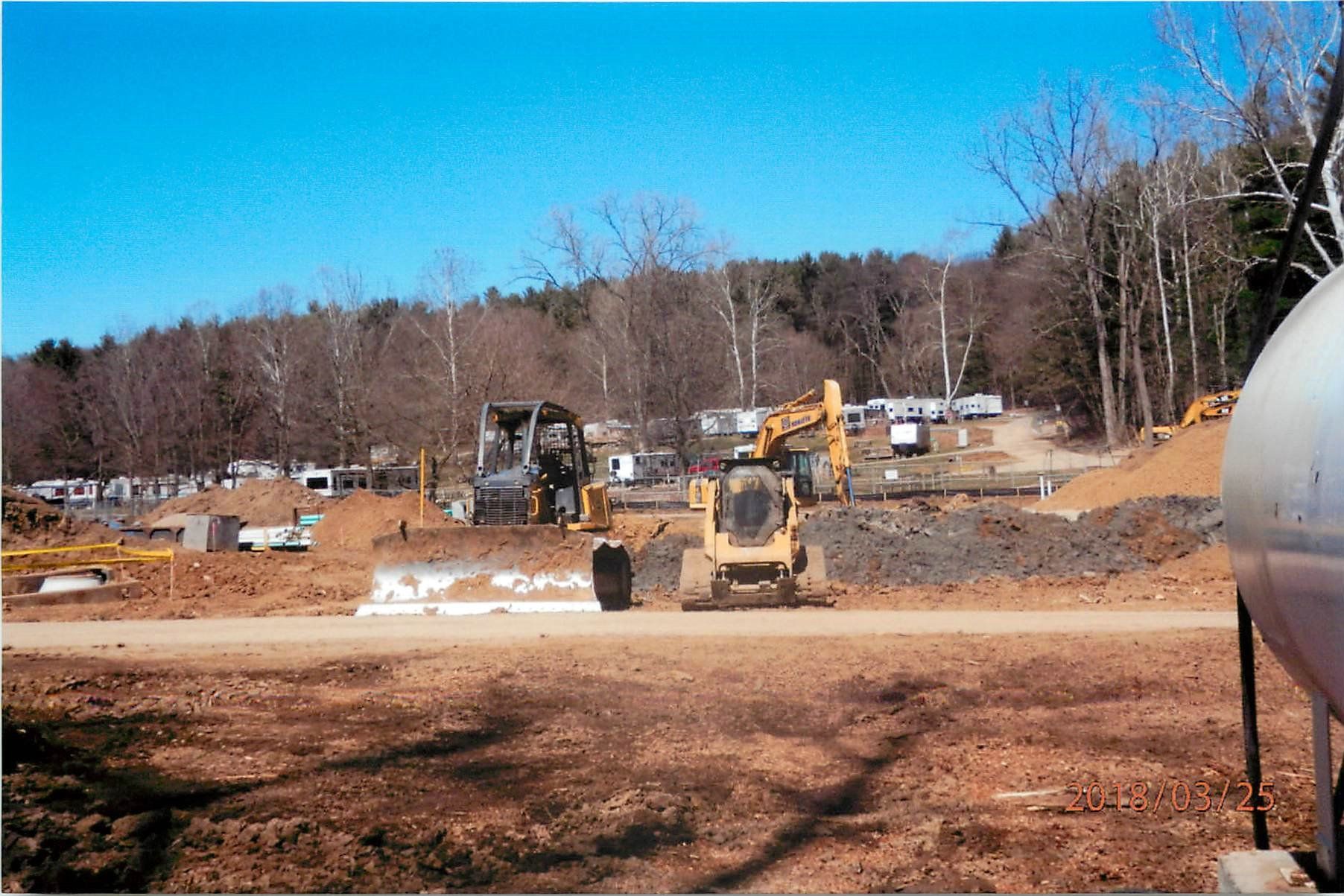 Construction site with heavy machinery, brown dirt, trees, and clear blue sky.