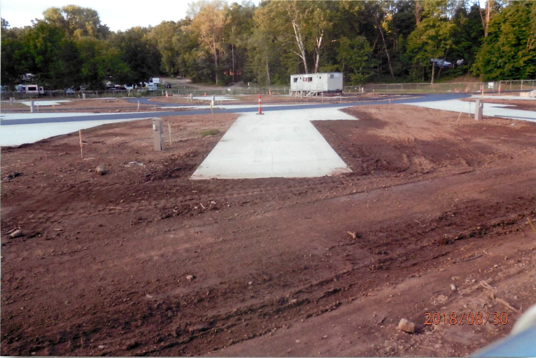 Dirt lot with concrete pads, possibly a construction site. Trees in background, small white building.