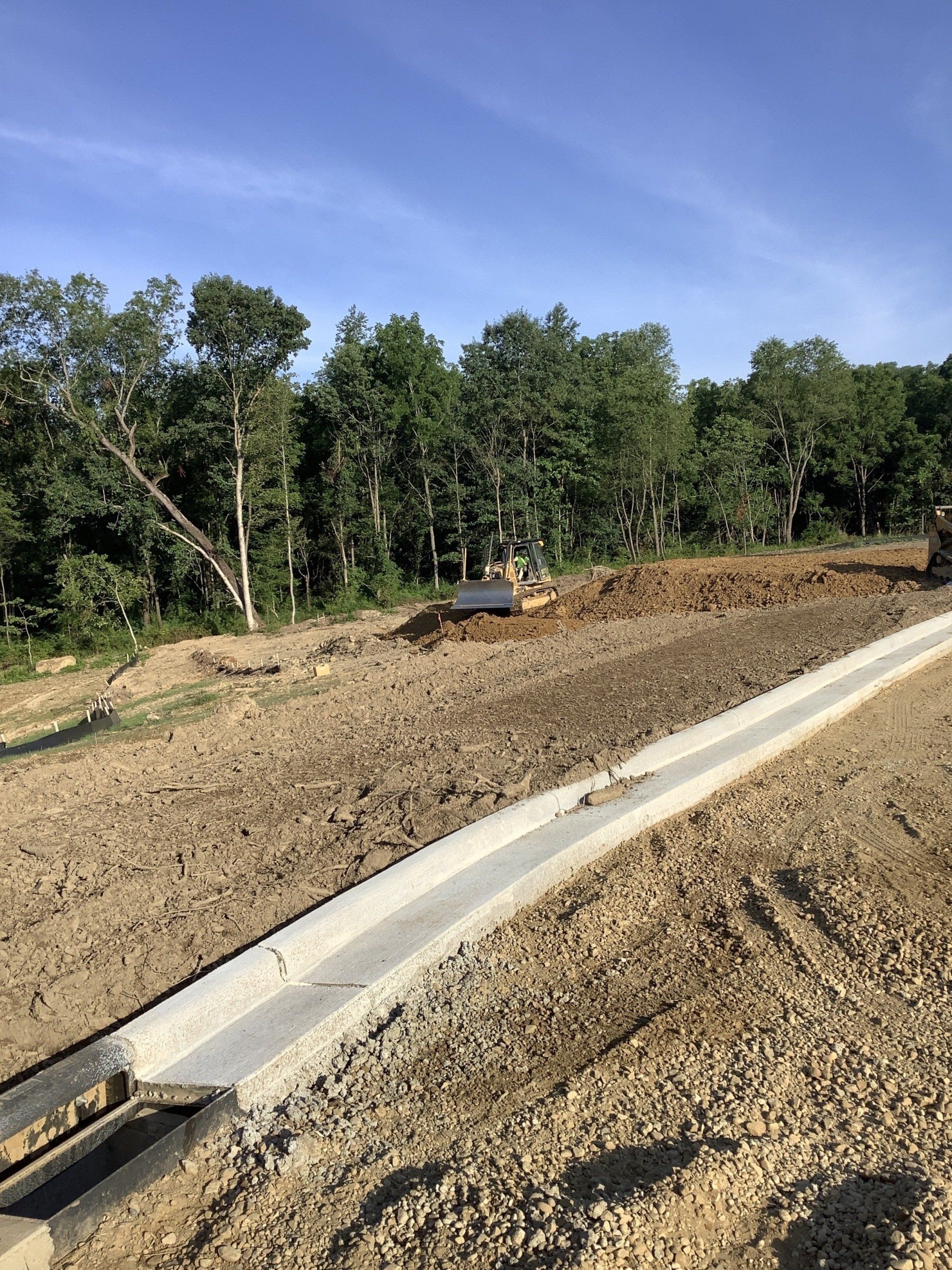 Construction site with a concrete drainage channel in foreground, bulldozer in the distance, and trees under a blue sky.