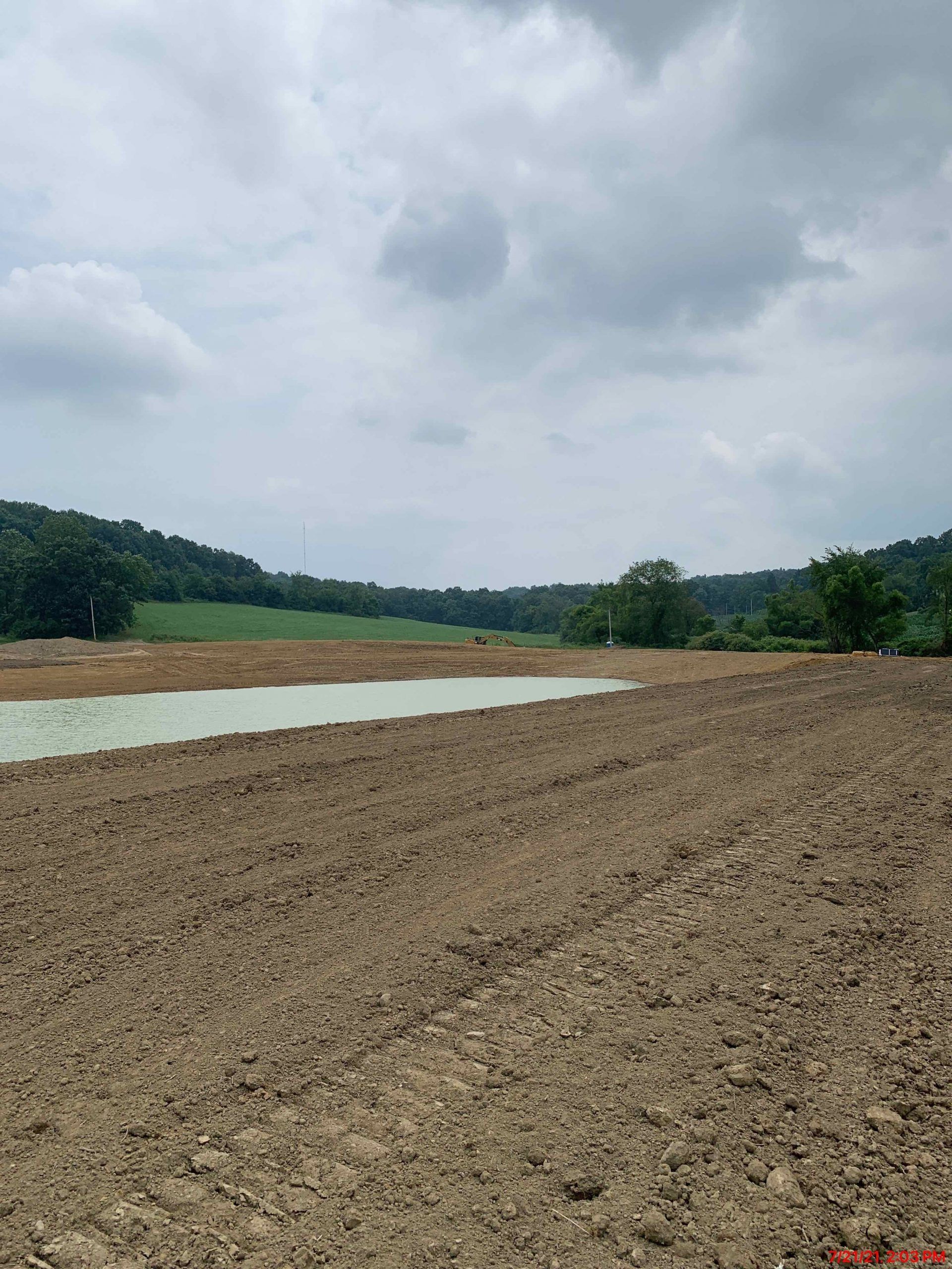 Plowed brown field with a green, water-filled area, surrounded by green trees under a cloudy sky.