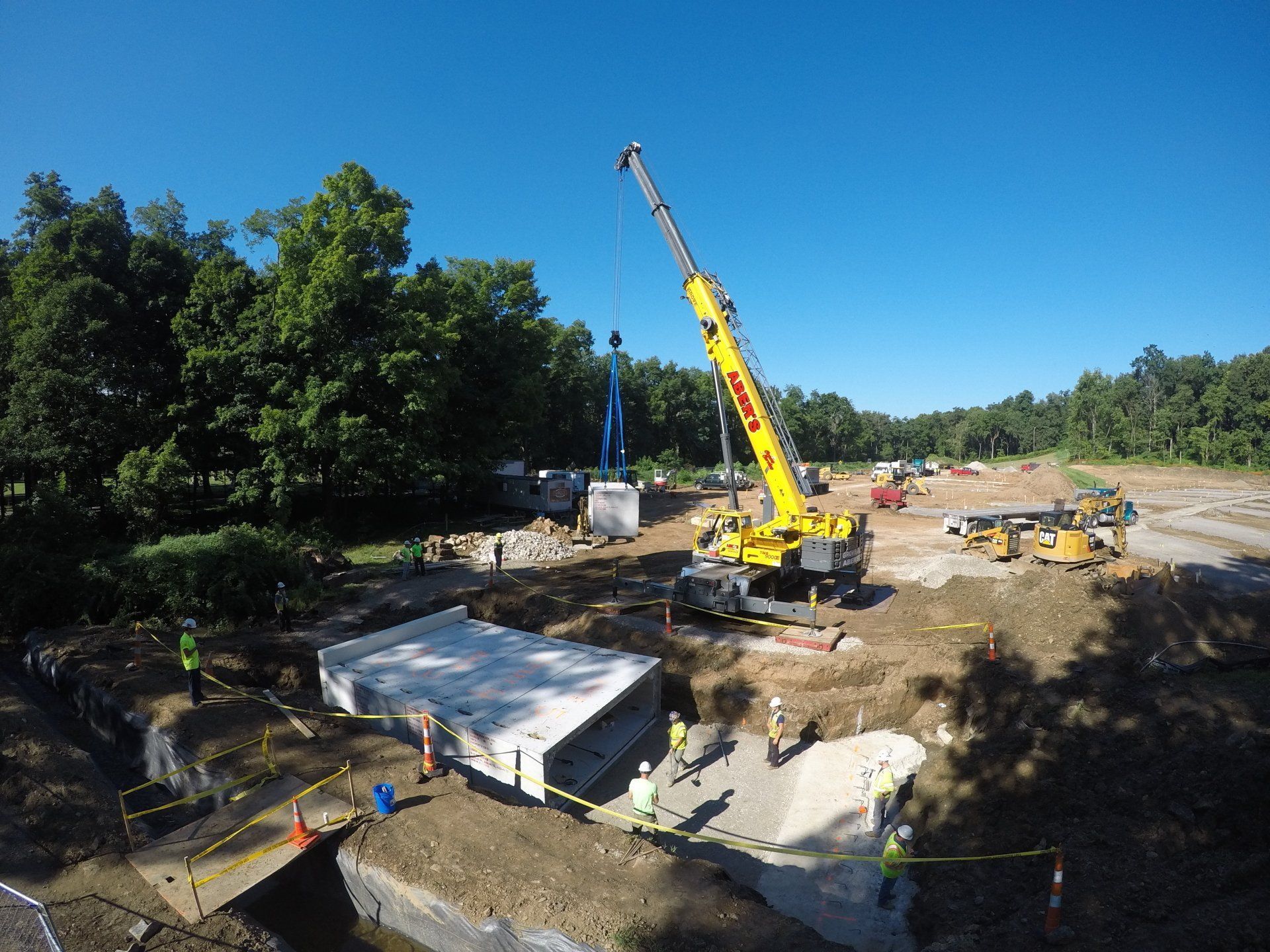 Crane lifting concrete bridge section at construction site. Workers, earth-moving equipment, trees.
