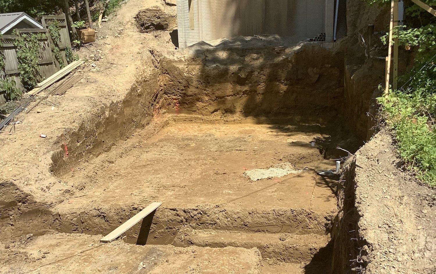 Excavation of a rectangular area in a yard, showing exposed dirt. A concrete structure is visible in the background.