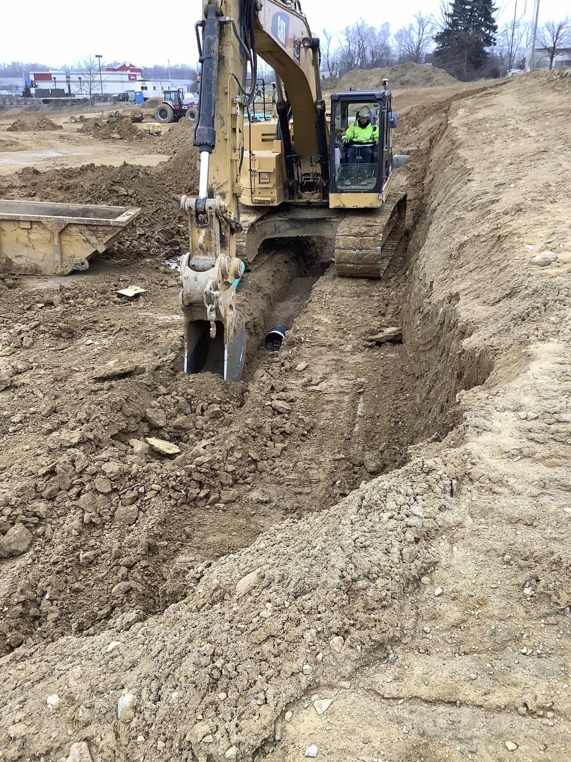 Excavator digging a trench on a construction site; operator in the cab, brown soil.