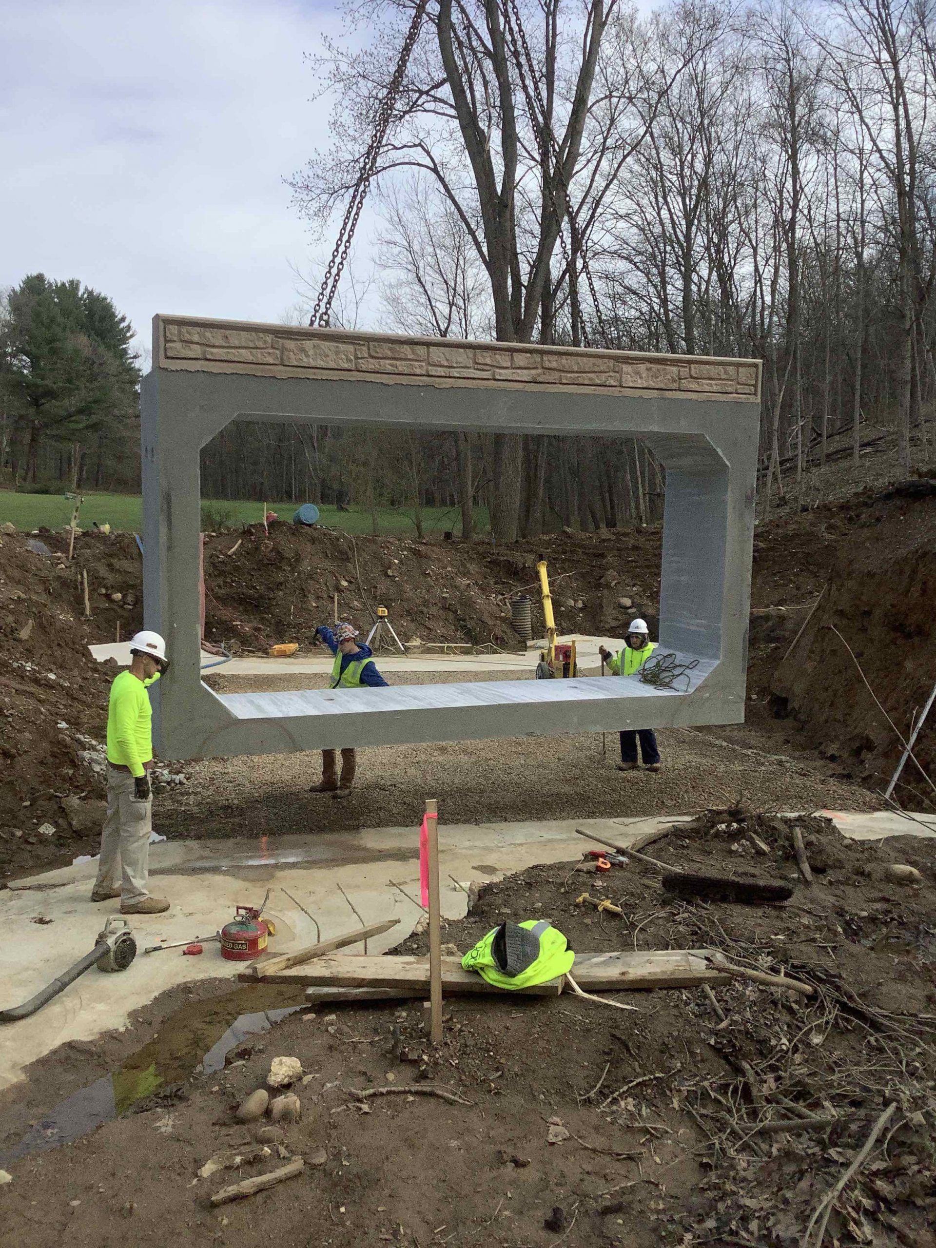 Construction workers lift a large concrete box structure. Outdoor setting with dirt, trees, and cloudy sky.