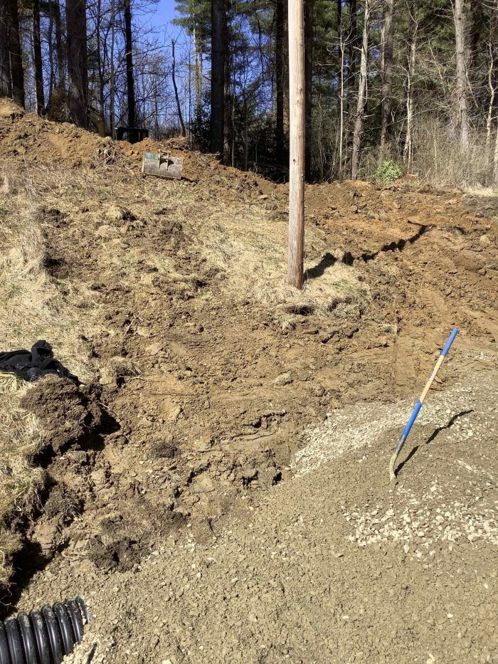 Earthen slope with utility pole. Dirt and gravel ground. Shovel leans against gravel. Wooded backdrop.