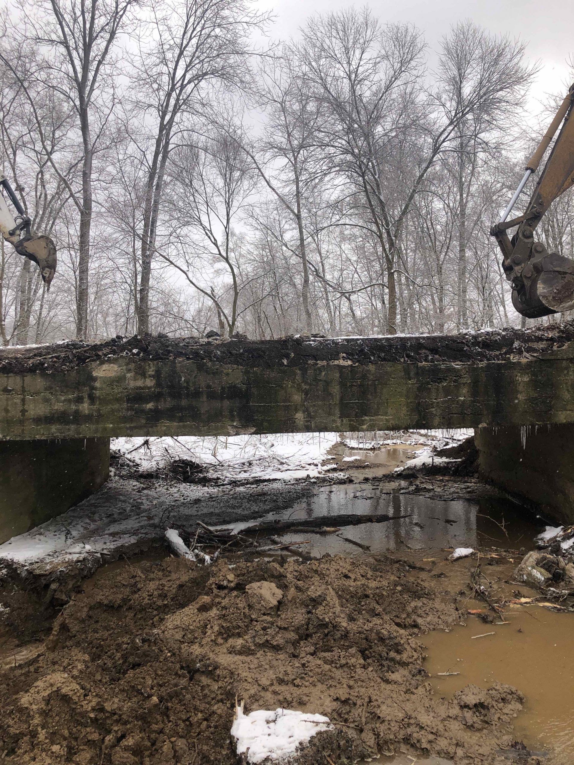 Construction site: A dilapidated bridge with an excavator. Muddy water and snow in foreground, trees in background.