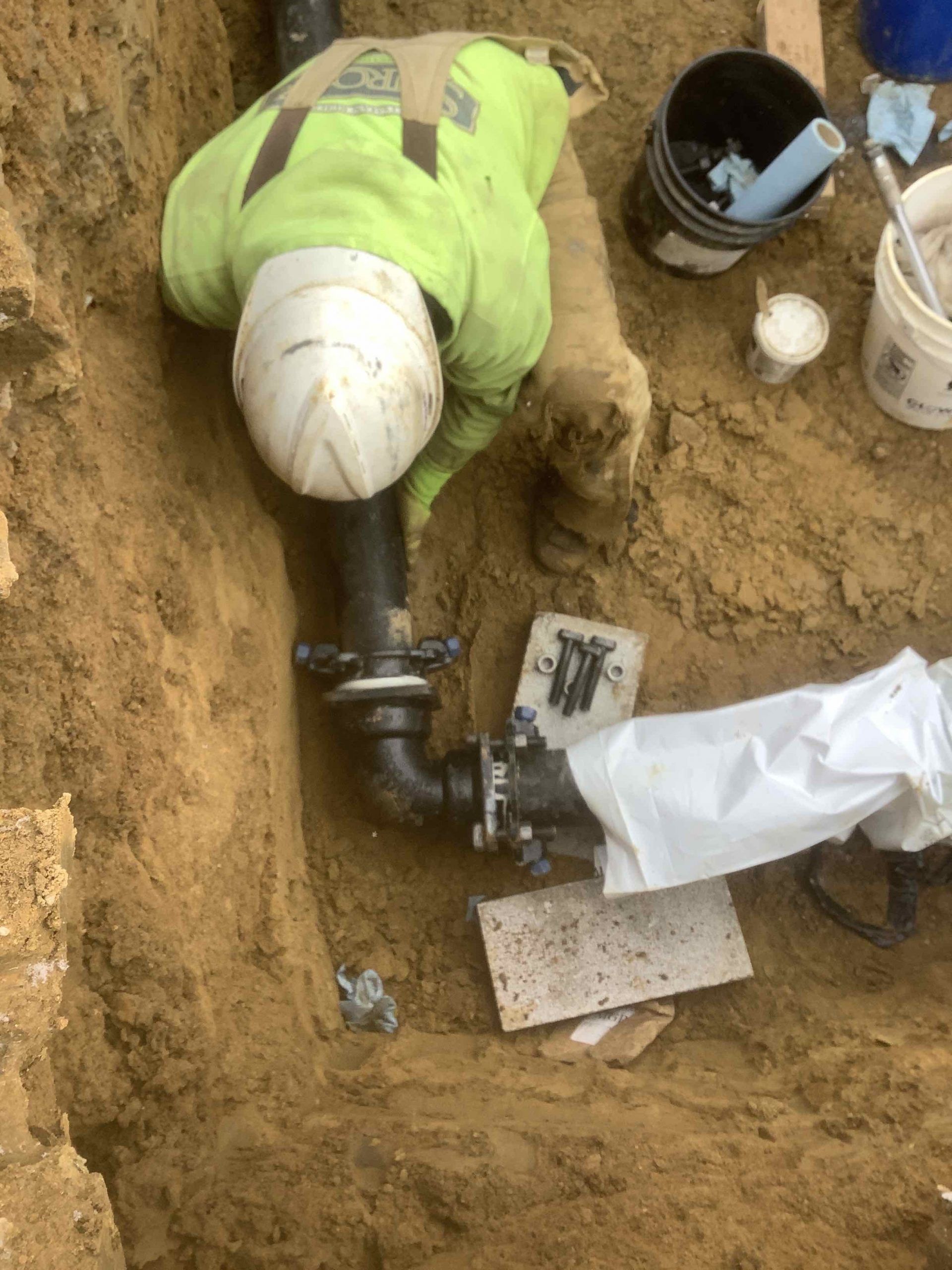 Worker in neon green jacket and white hard hat installing pipes in a trench.