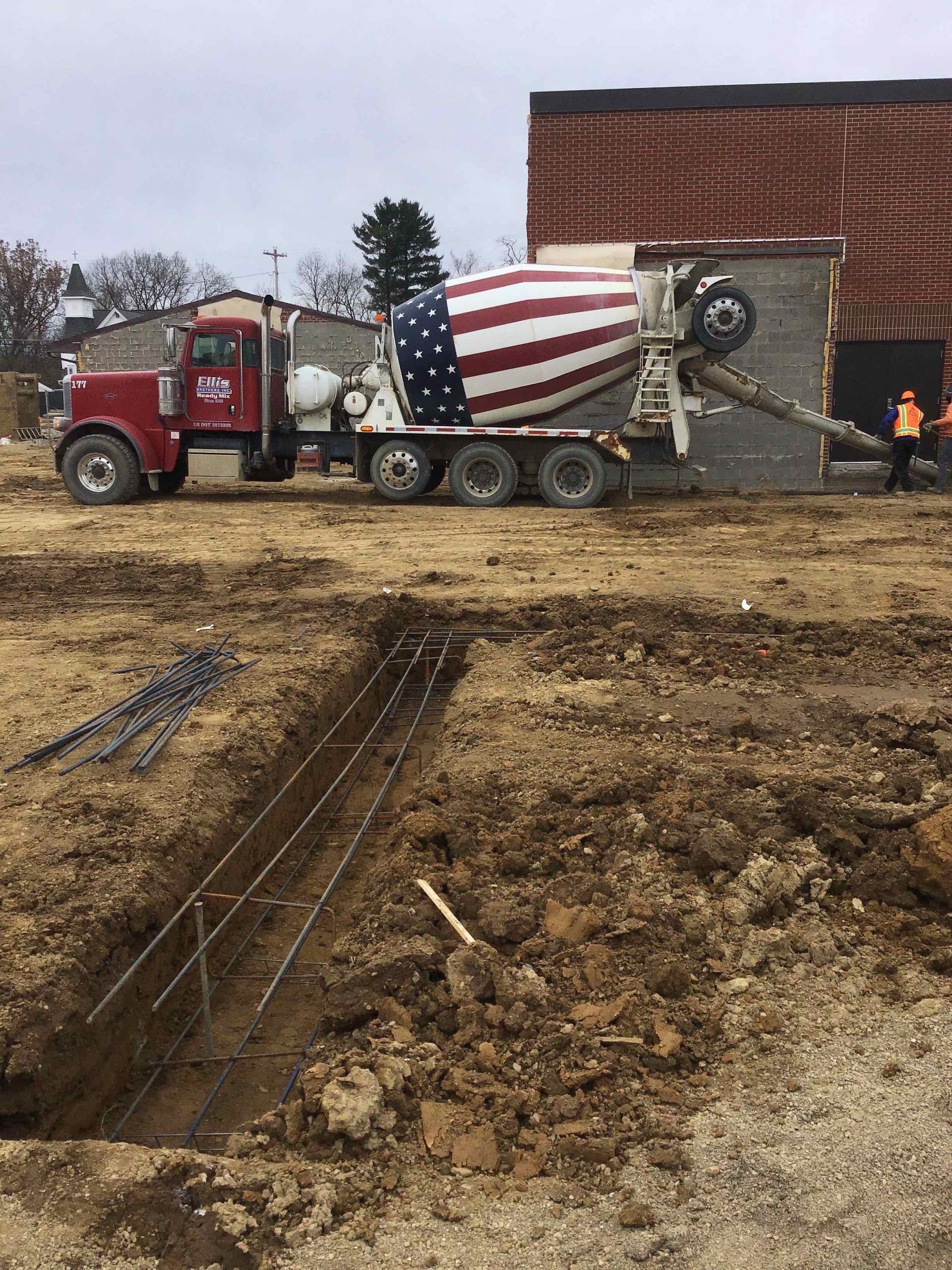 Red and white cement truck with American flag design pouring concrete at a construction site.
