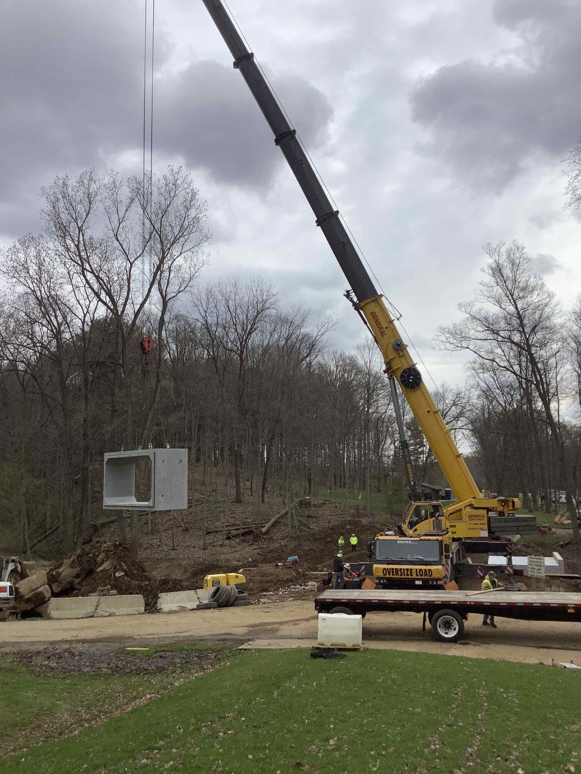 Crane lifting a metal structure at a construction site with trees in the background. Cloudy day.