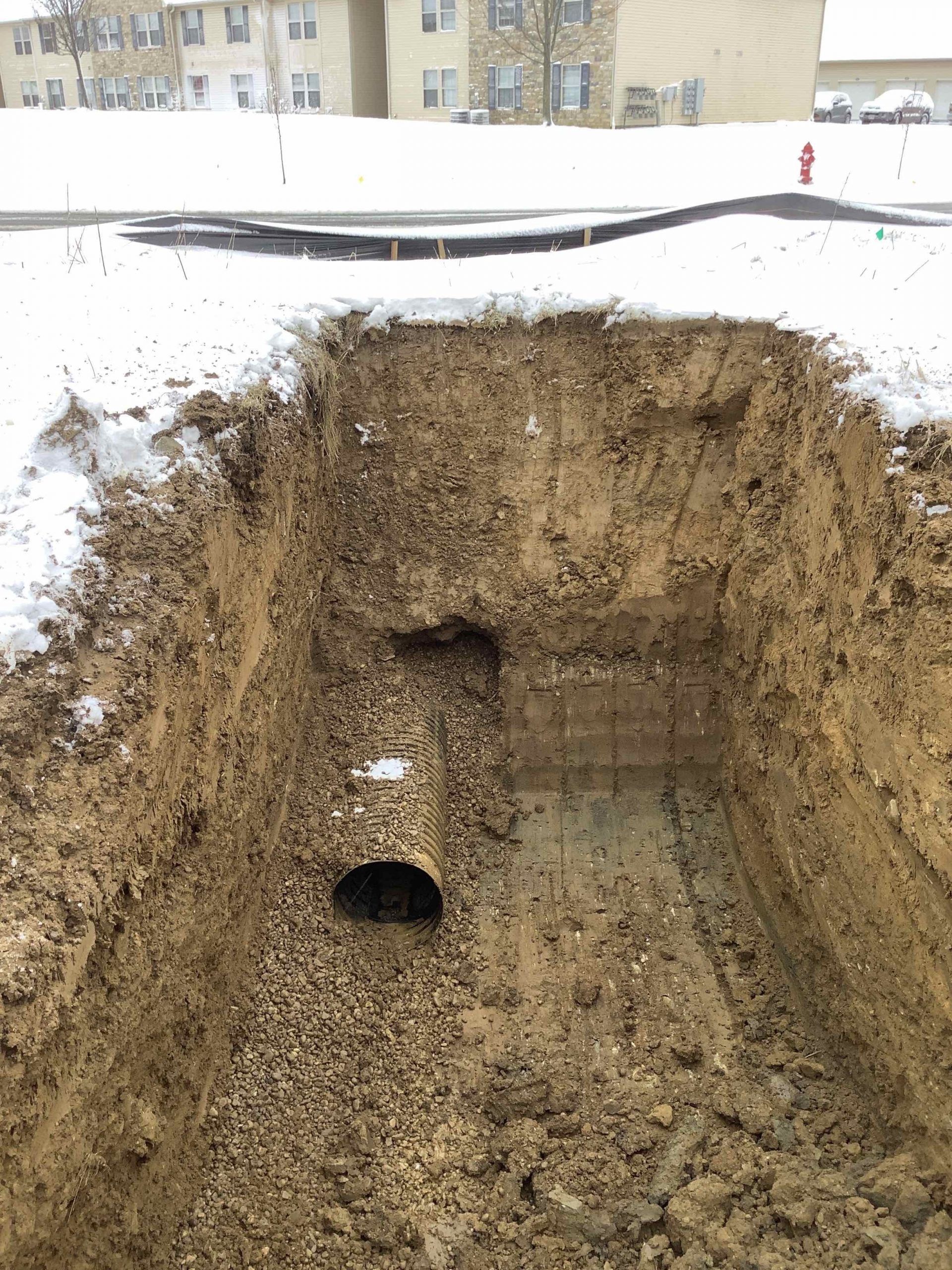 A trench dug in the snowy ground reveals a buried pipe; apartments in the background.