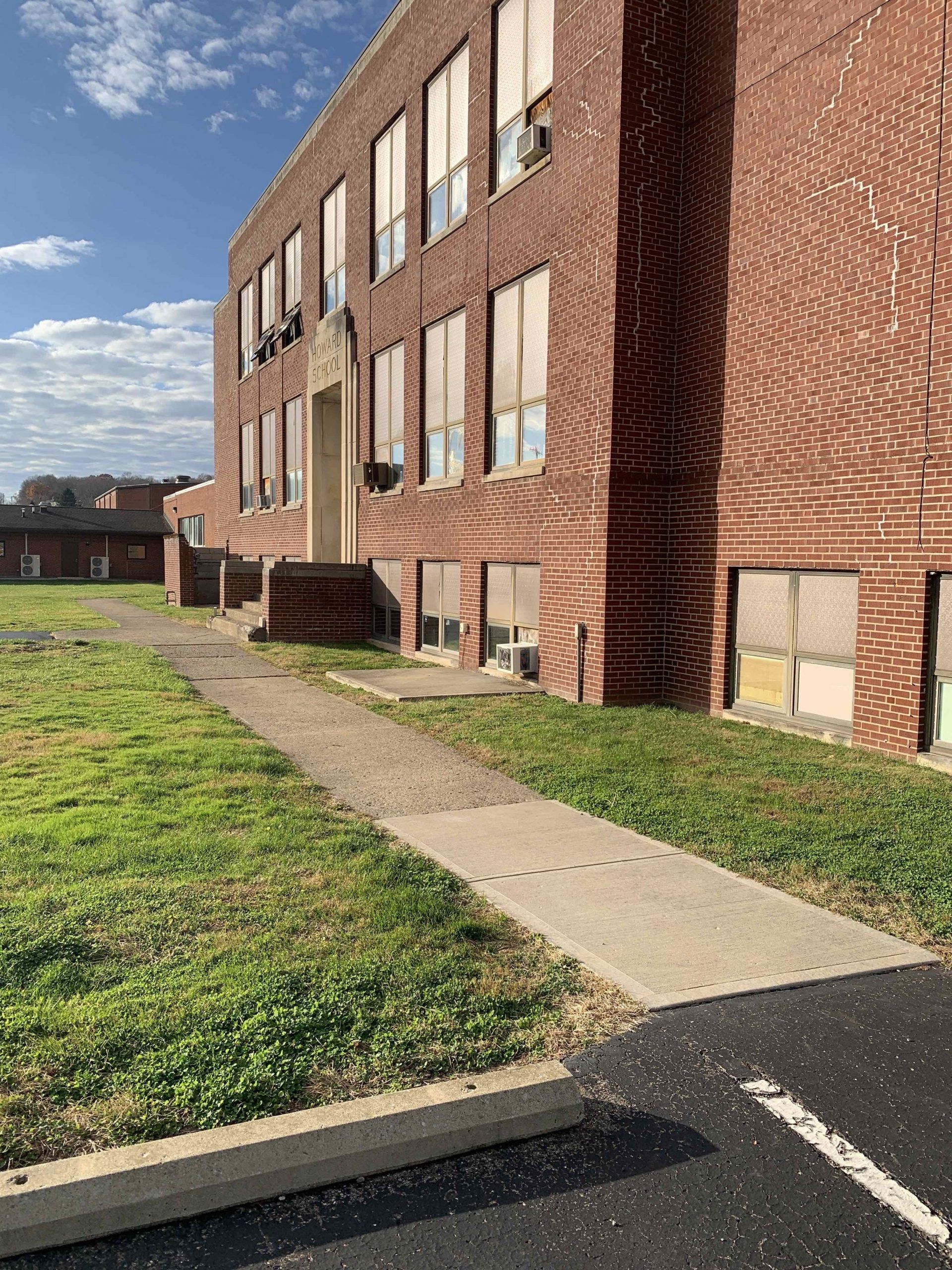 Brick building with boarded-up windows, overgrown grass, and a concrete walkway.