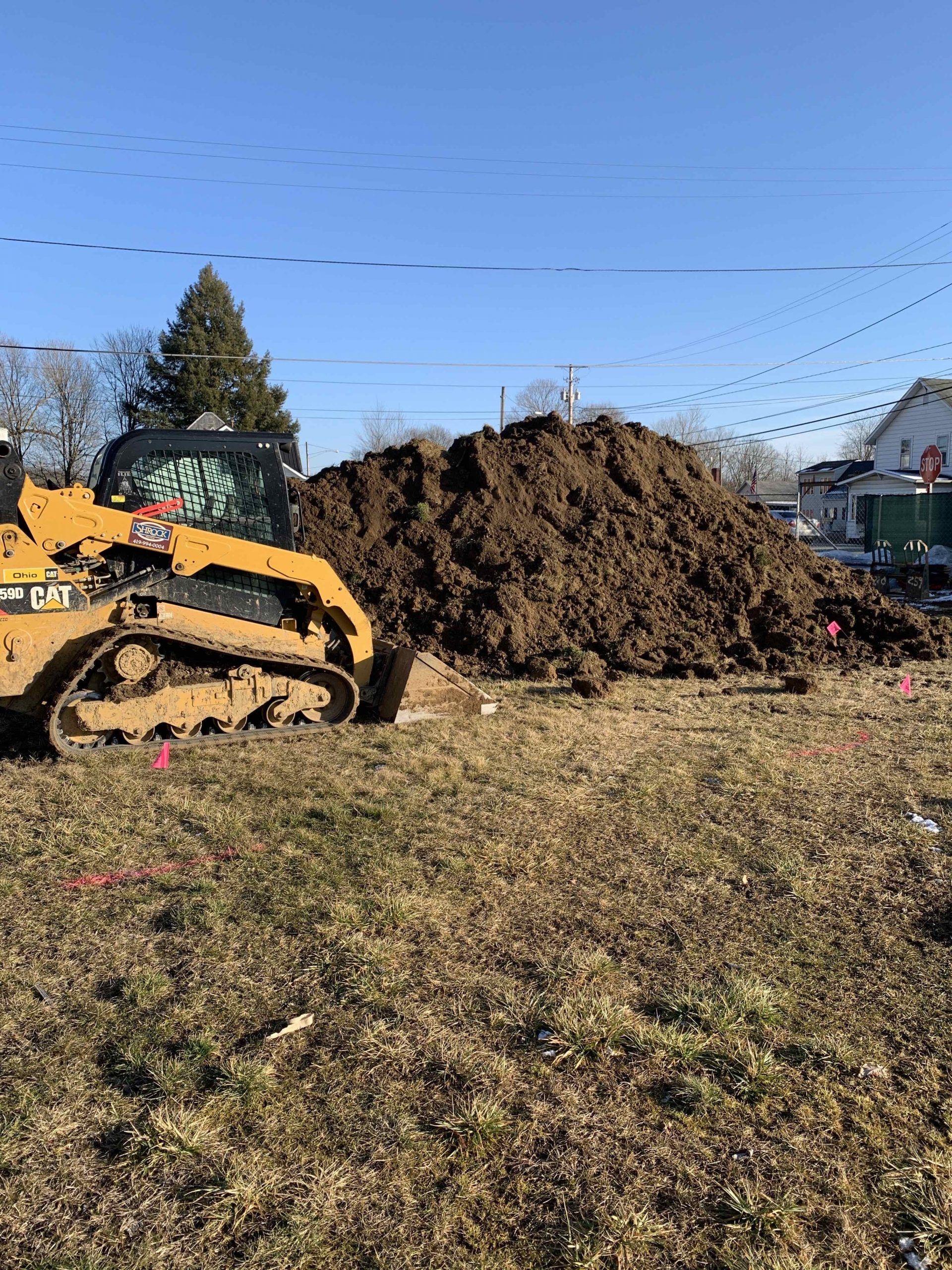 Yellow bulldozer next to a large pile of dirt in a grassy field. Power lines and buildings in the background.
