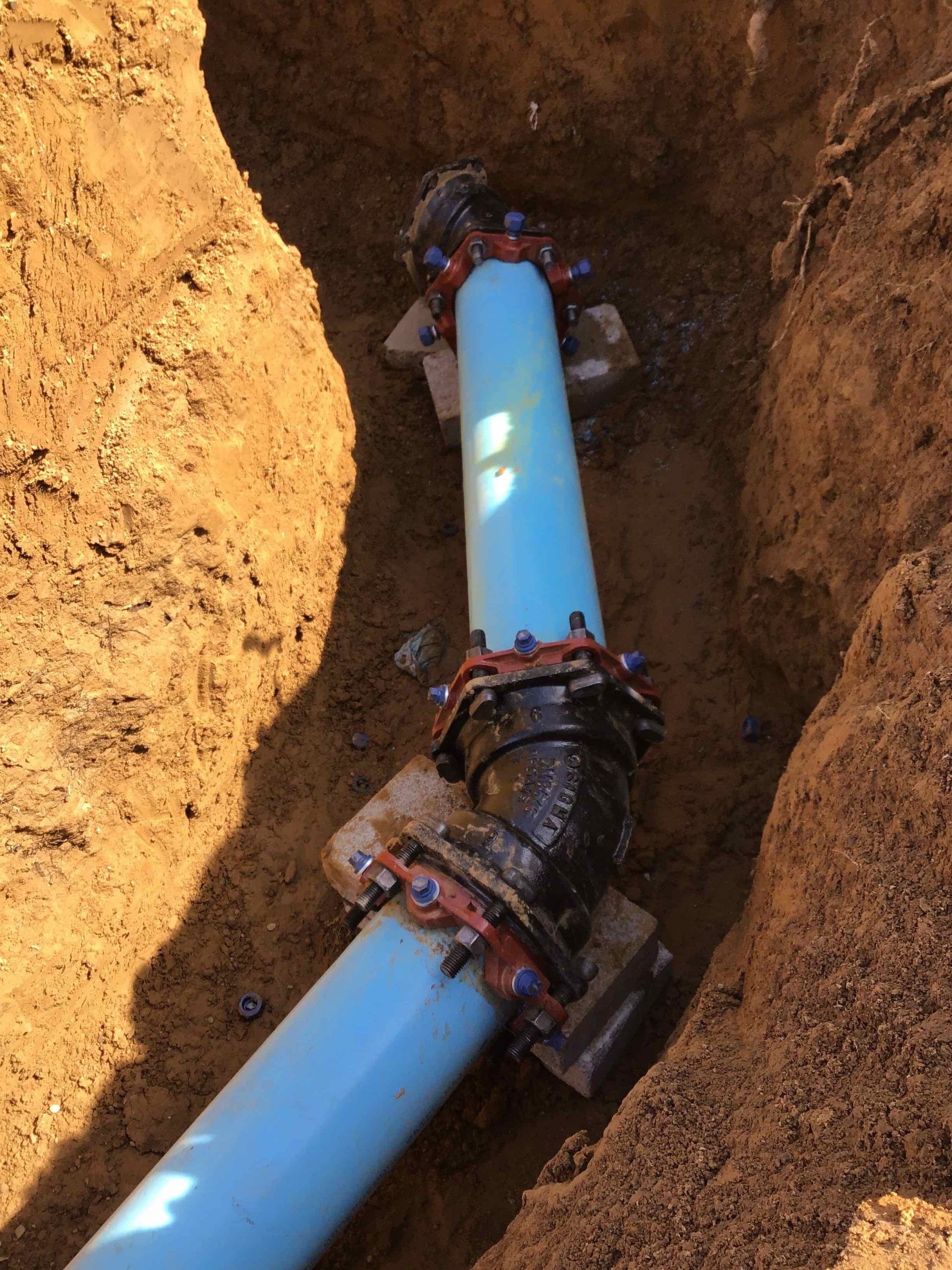 Blue and black pipes joined with bolted connectors in a dirt trench, supported by concrete blocks.