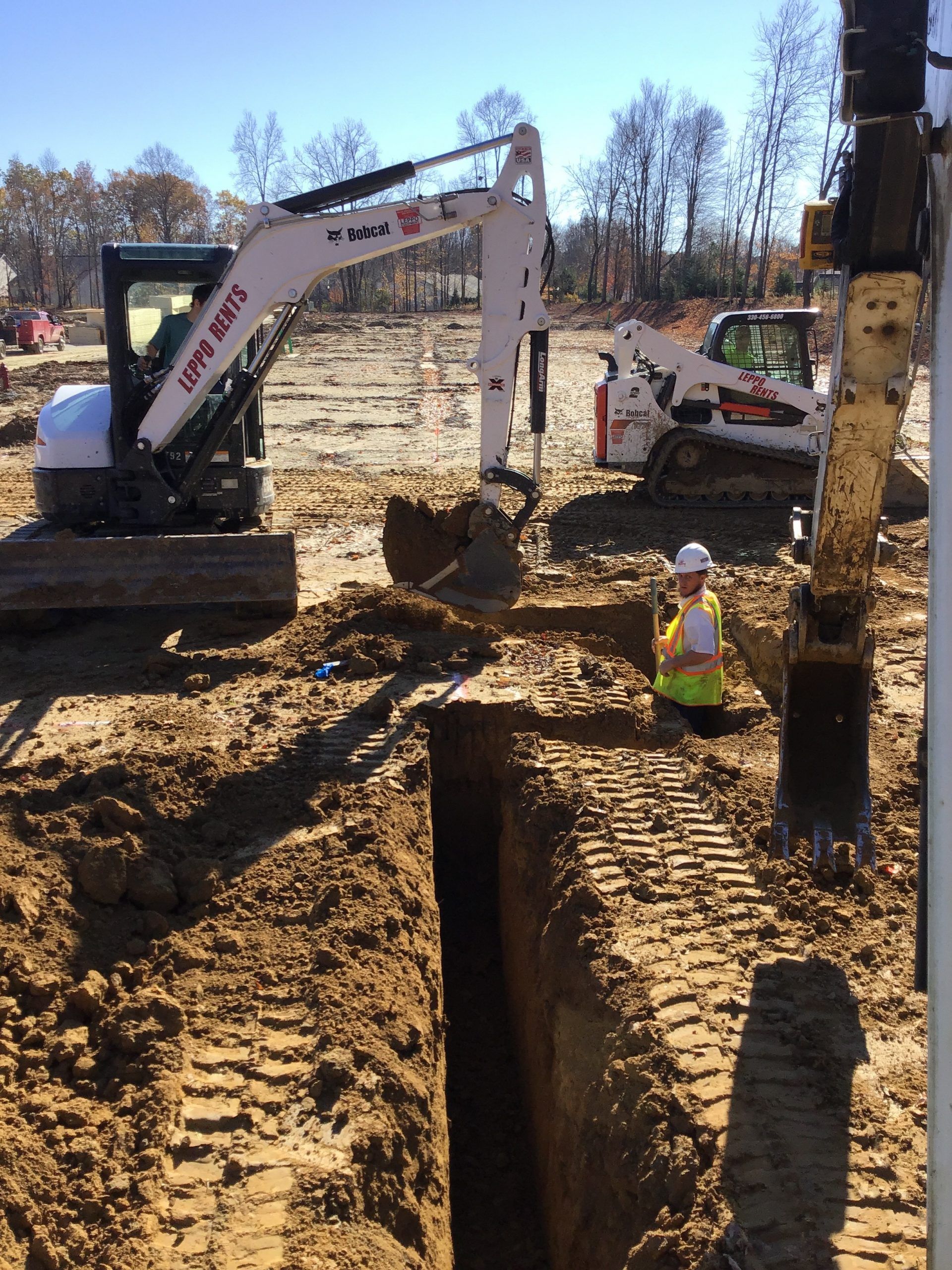 Construction site with an excavator digging a trench; worker in high-vis vest; a Bobcat and drill rig present.
