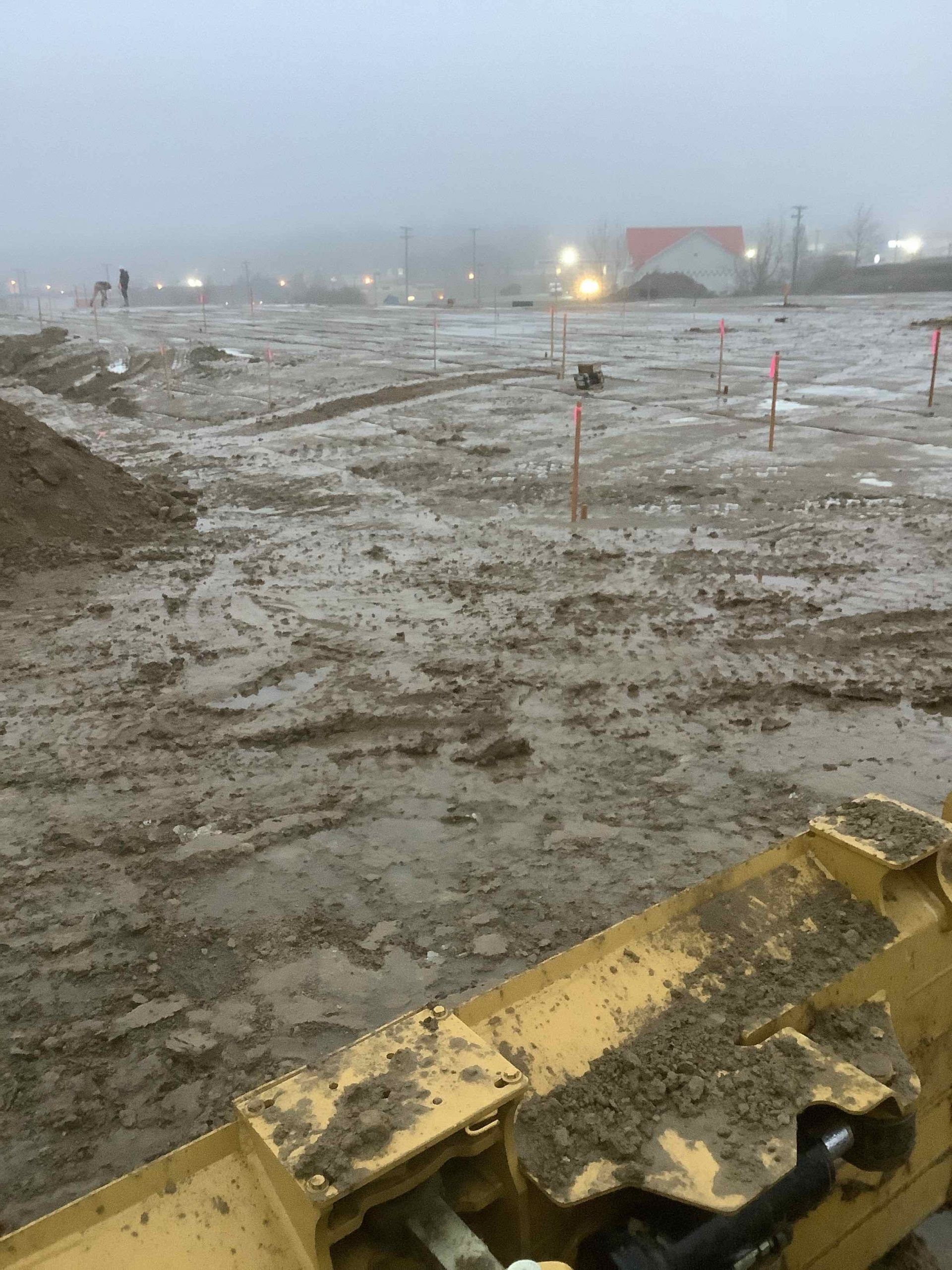 Muddy construction site under overcast sky with orange markers and heavy equipment in the foreground.