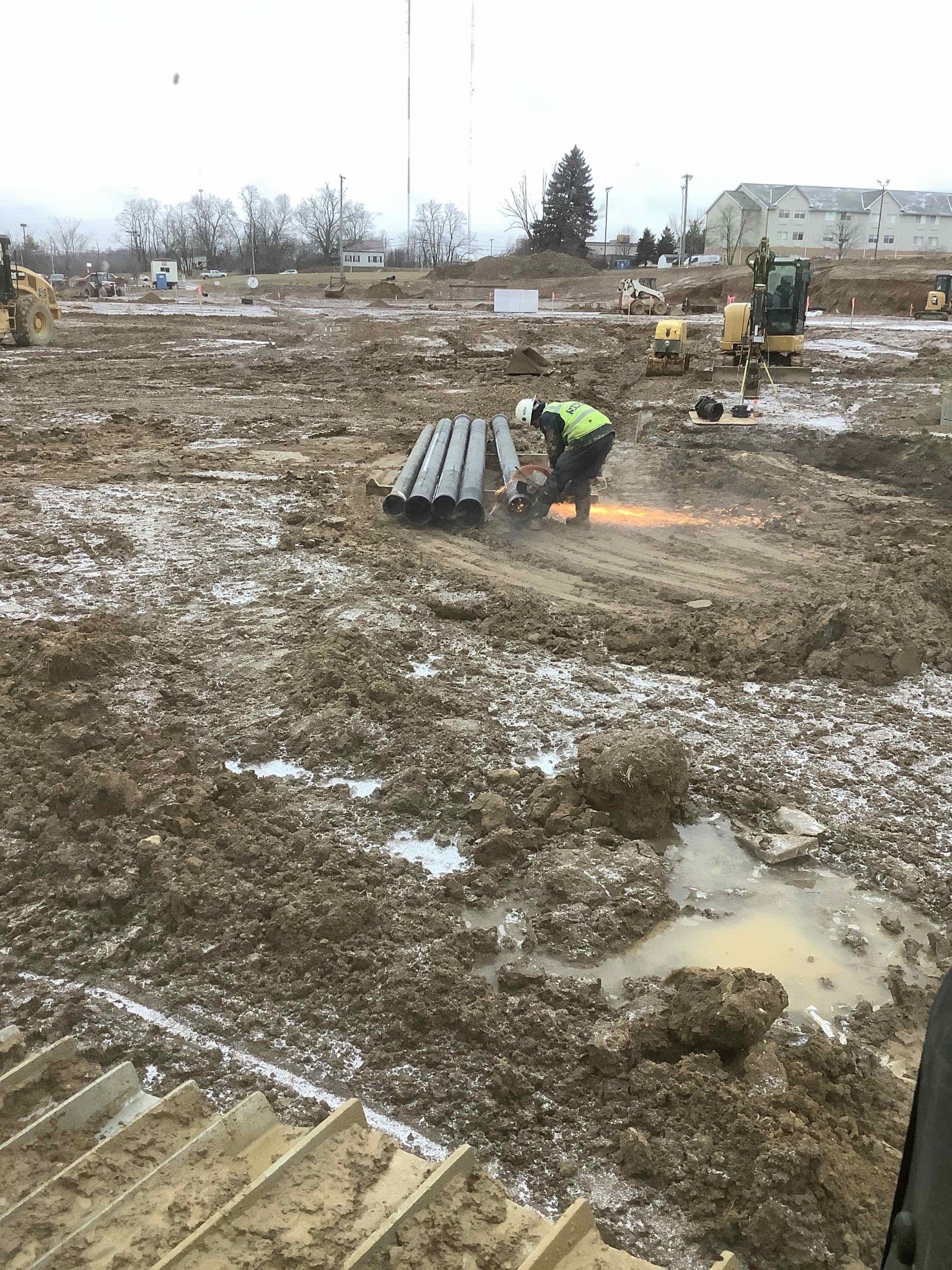 Construction worker cutting metal on muddy ground. Yellow excavator and buildings in the background. Overcast day.