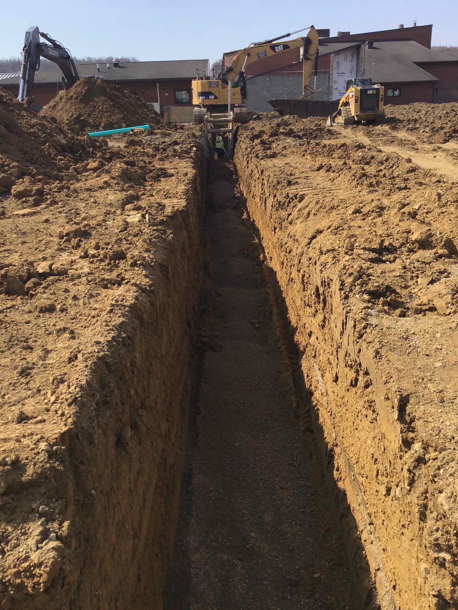 Construction site with a deep trench in brown dirt, machinery in the background.