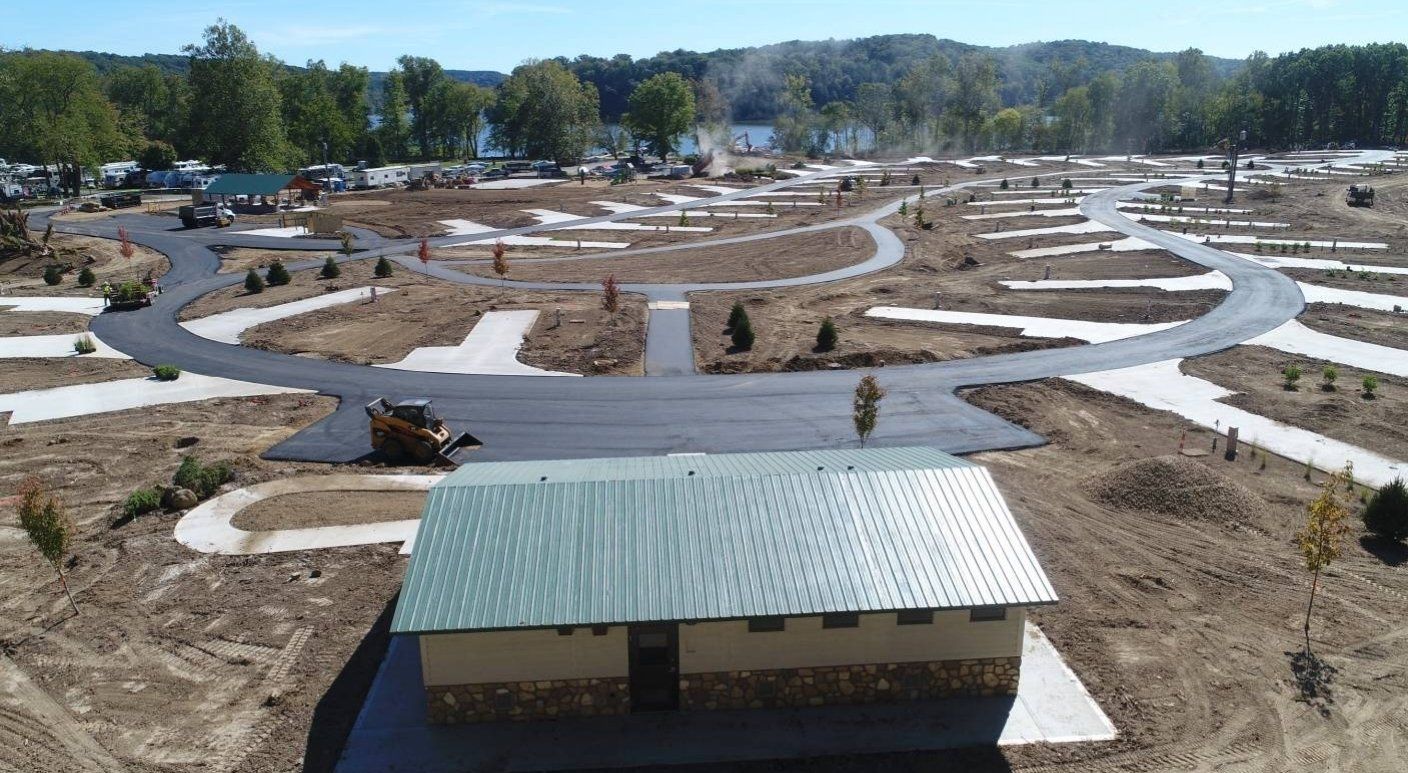 Aerial view of a campsite with paved roads, concrete pads, and a restroom building; dirt and new plantings surround.