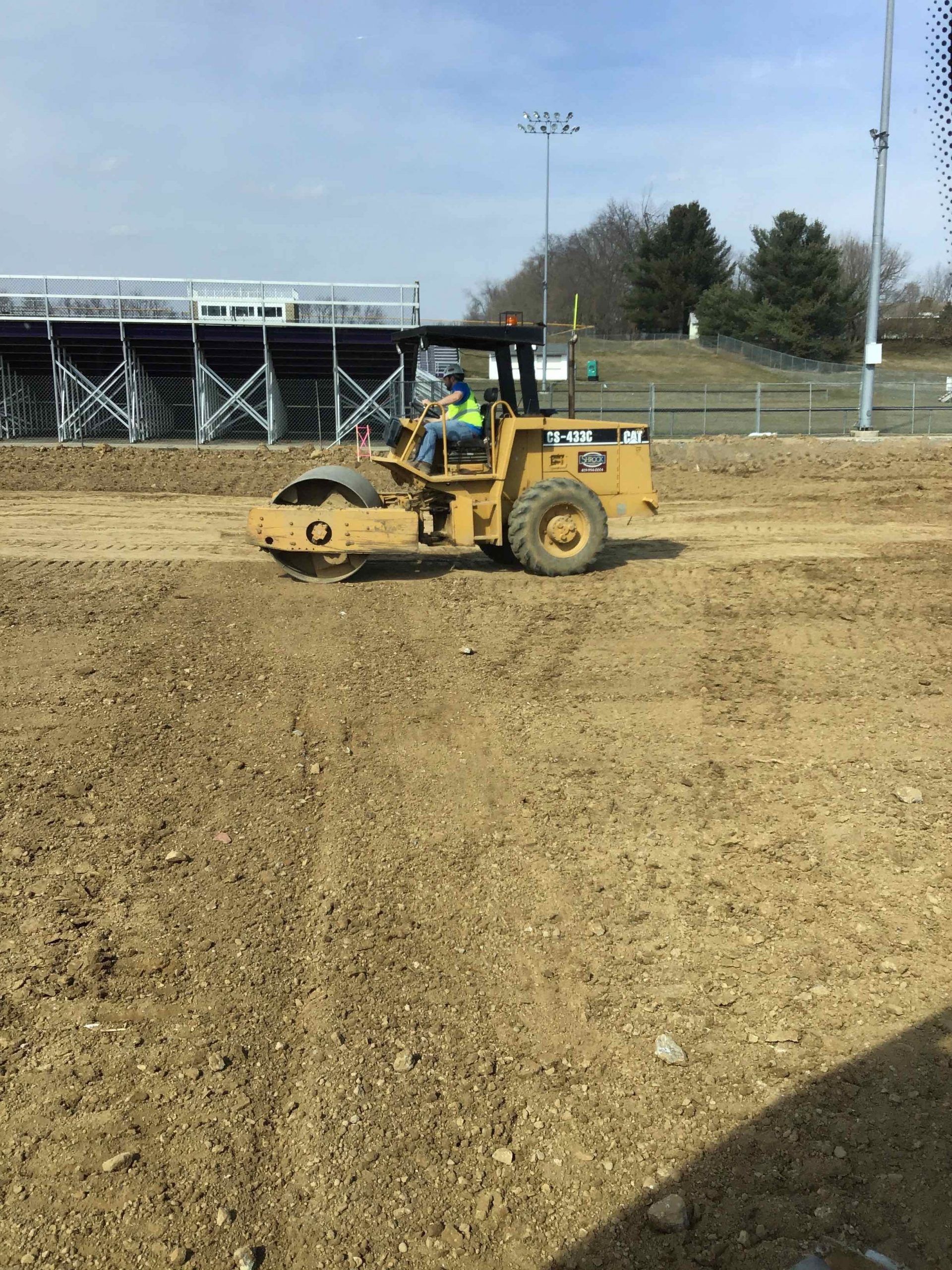 A yellow Caterpillar compactor machine rolls over dirt at a construction site near bleachers under a blue sky.