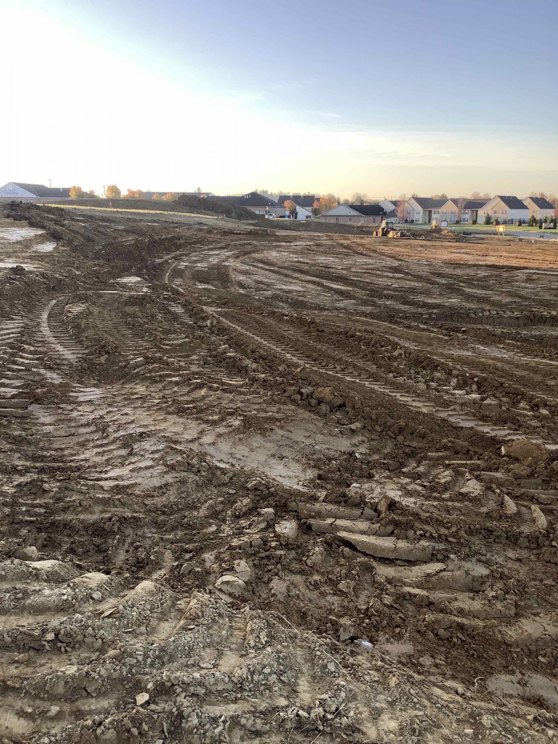 Muddy construction site with tire tracks, distant houses under a blue sky.