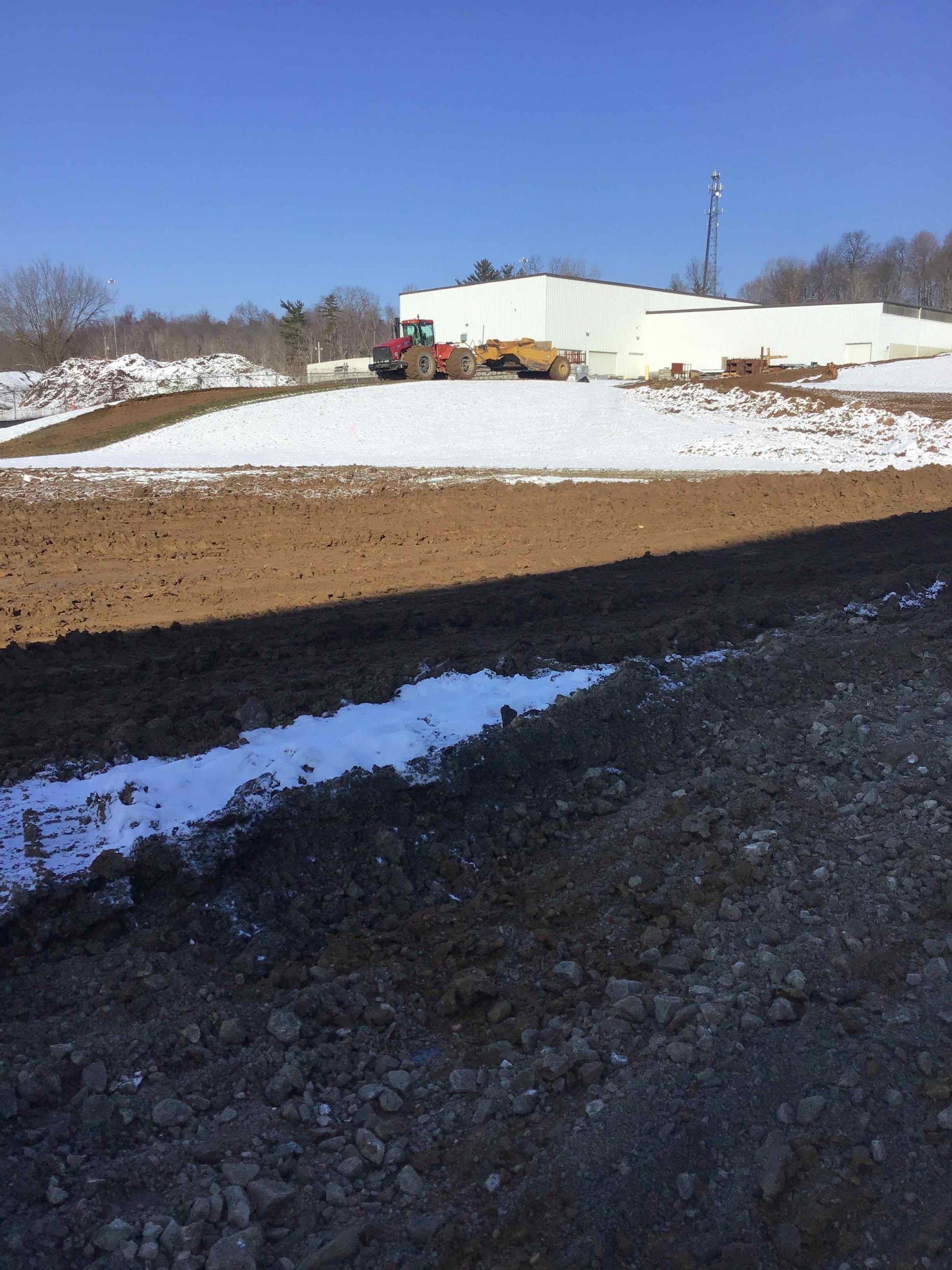 Construction site with snow on the ground; a tractor and excavator in front of a white building on a sunny day.