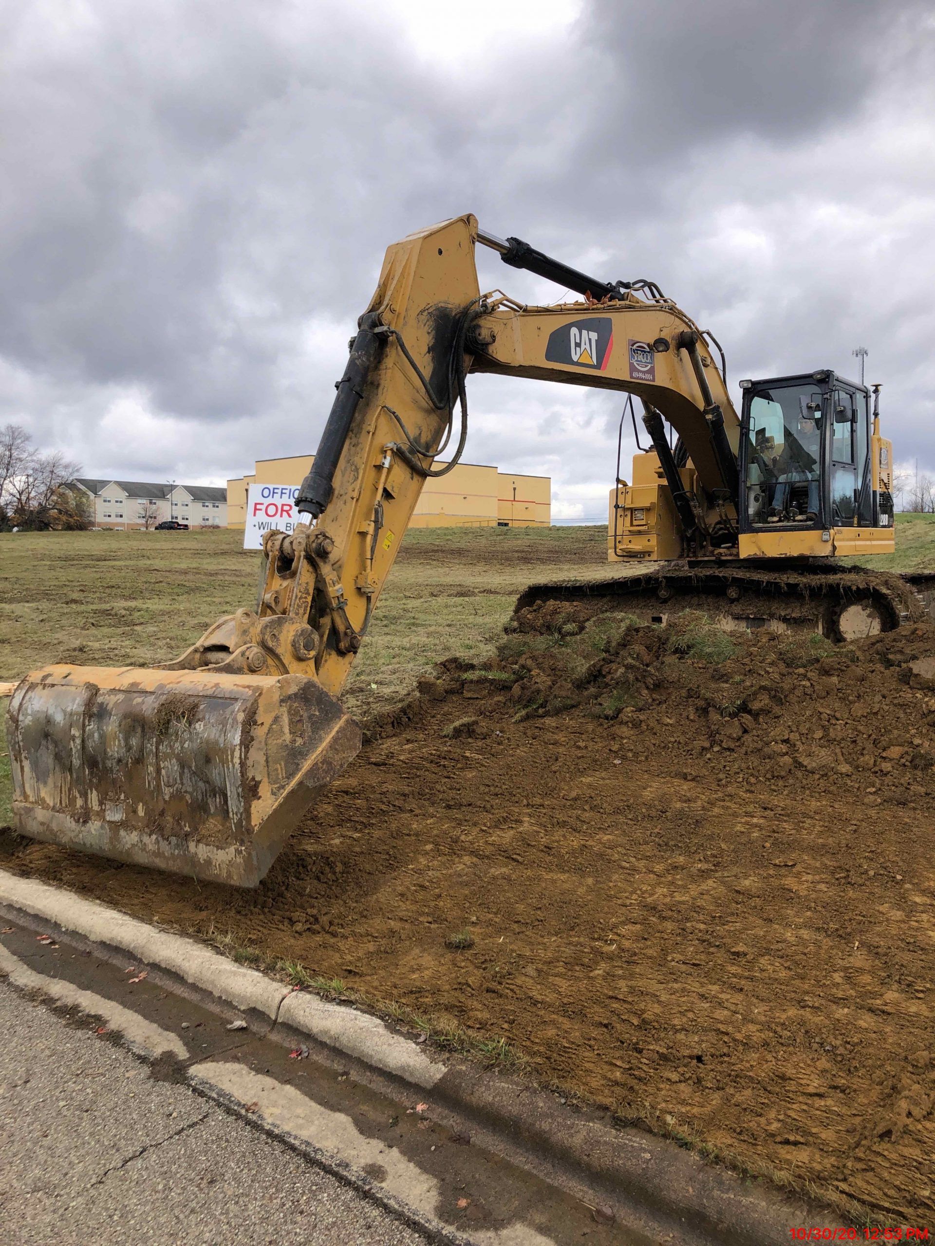 Yellow Caterpillar excavator digging in a grassy field next to a paved road. Overcast sky.