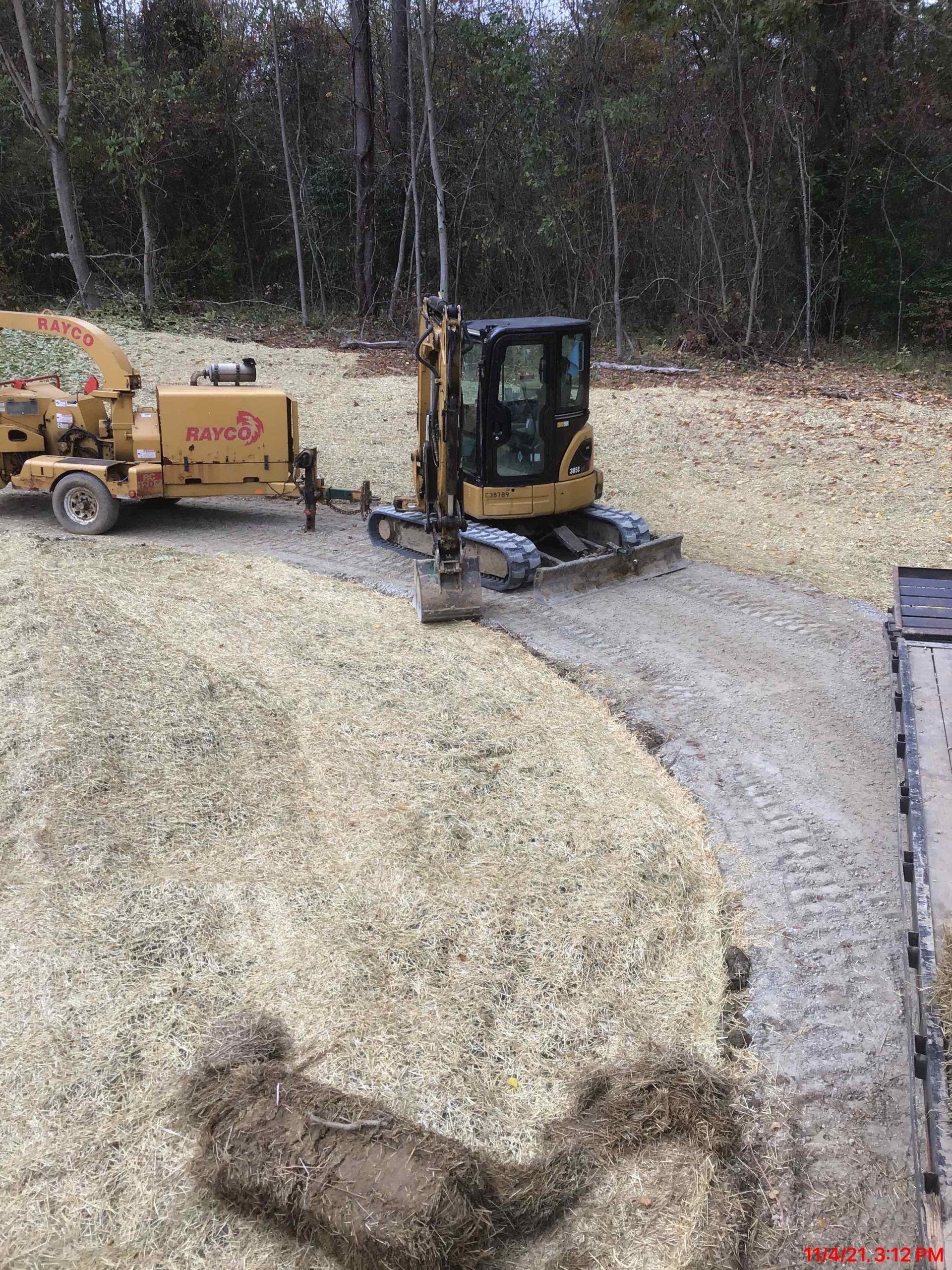 Mini excavator working on a wood-chipped area next to a trailer-mounted chipper. Forest background.