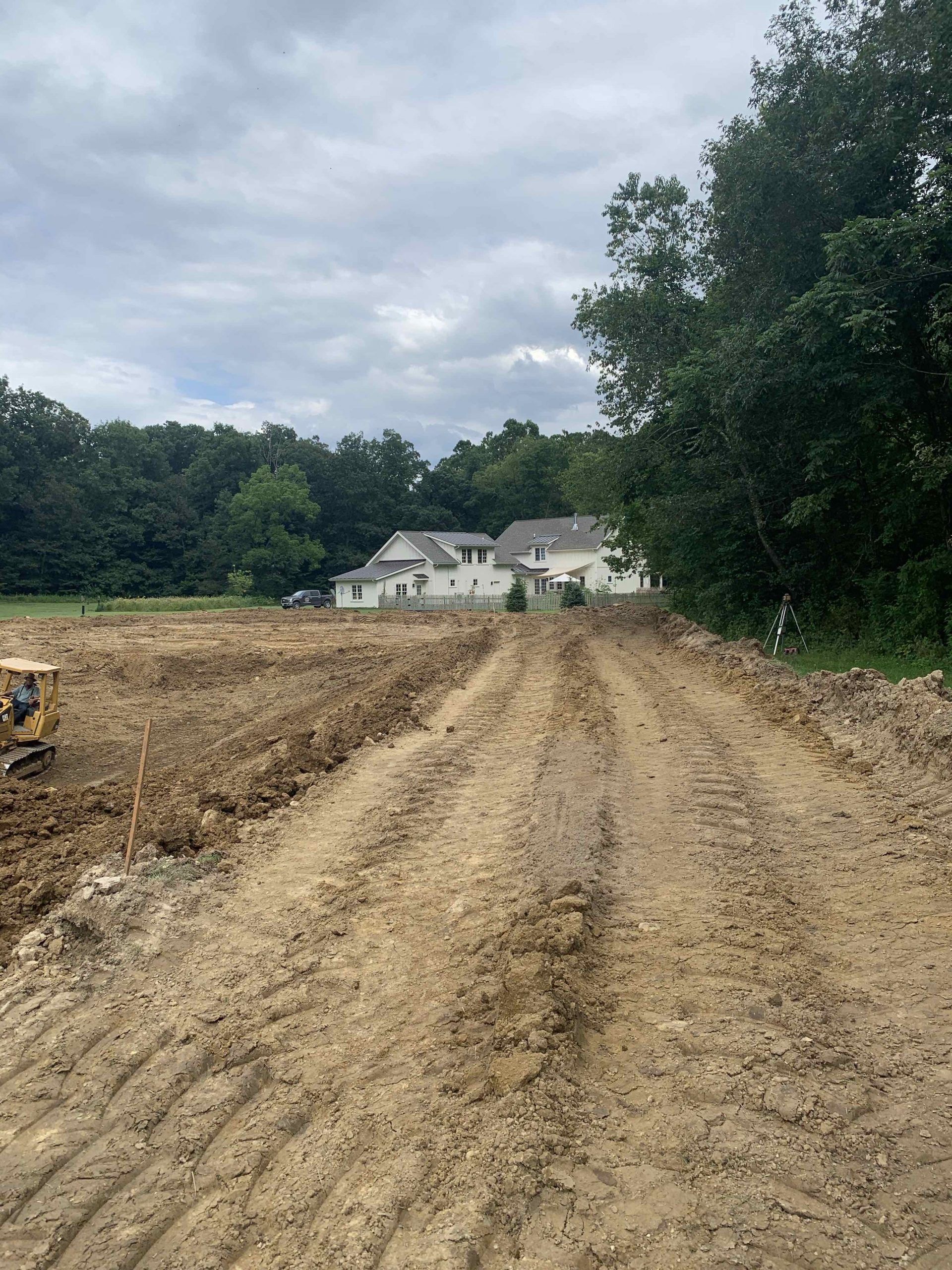 Dirt road through a construction site, leading to a house in the distance under a cloudy sky.