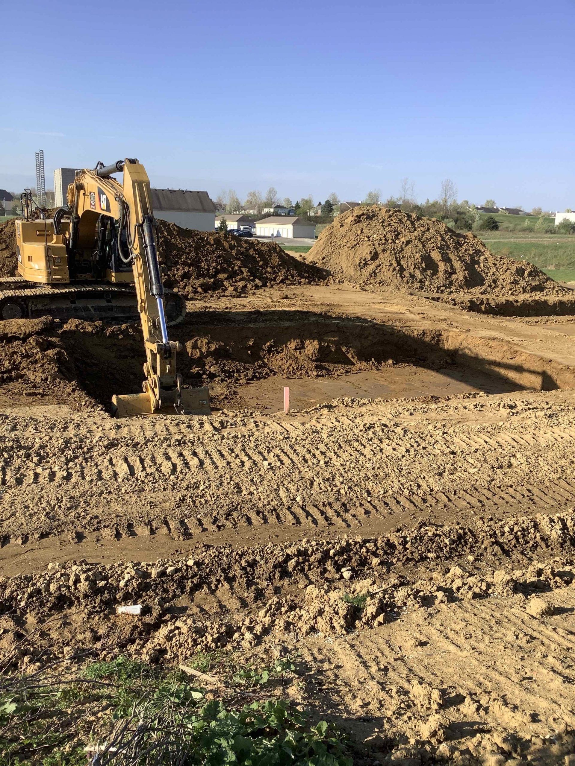 Excavator digging in dirt, with a pile of earth beside it. Sunny day, residential area in the background.