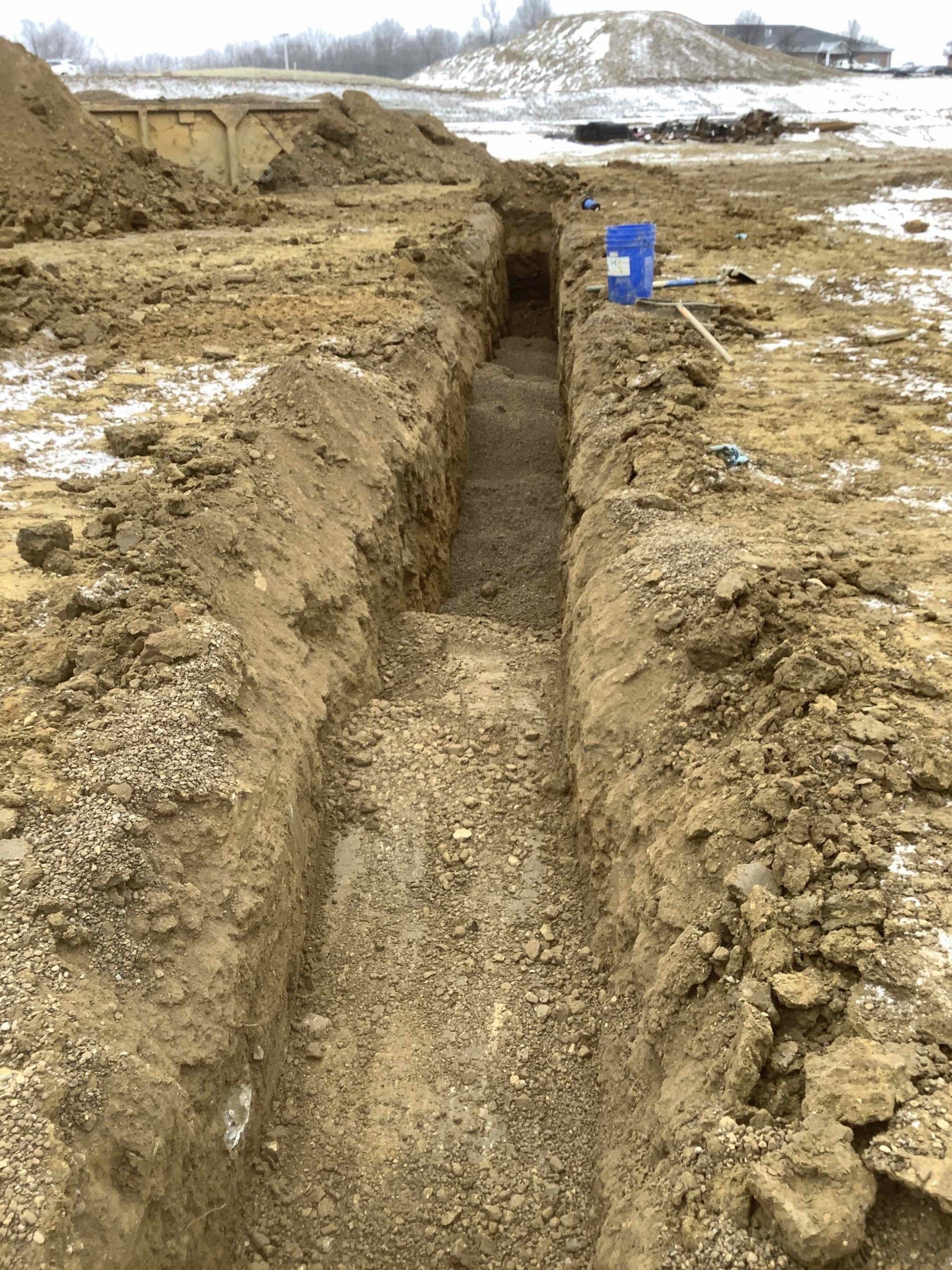 A trench dug in dirt on a construction site with a blue bucket and tools visible.