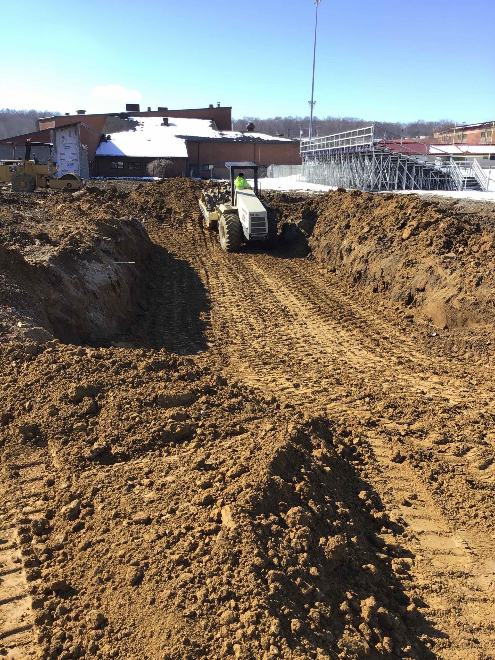 A tractor operating in a large dirt excavation pit; snowy buildings in the background.