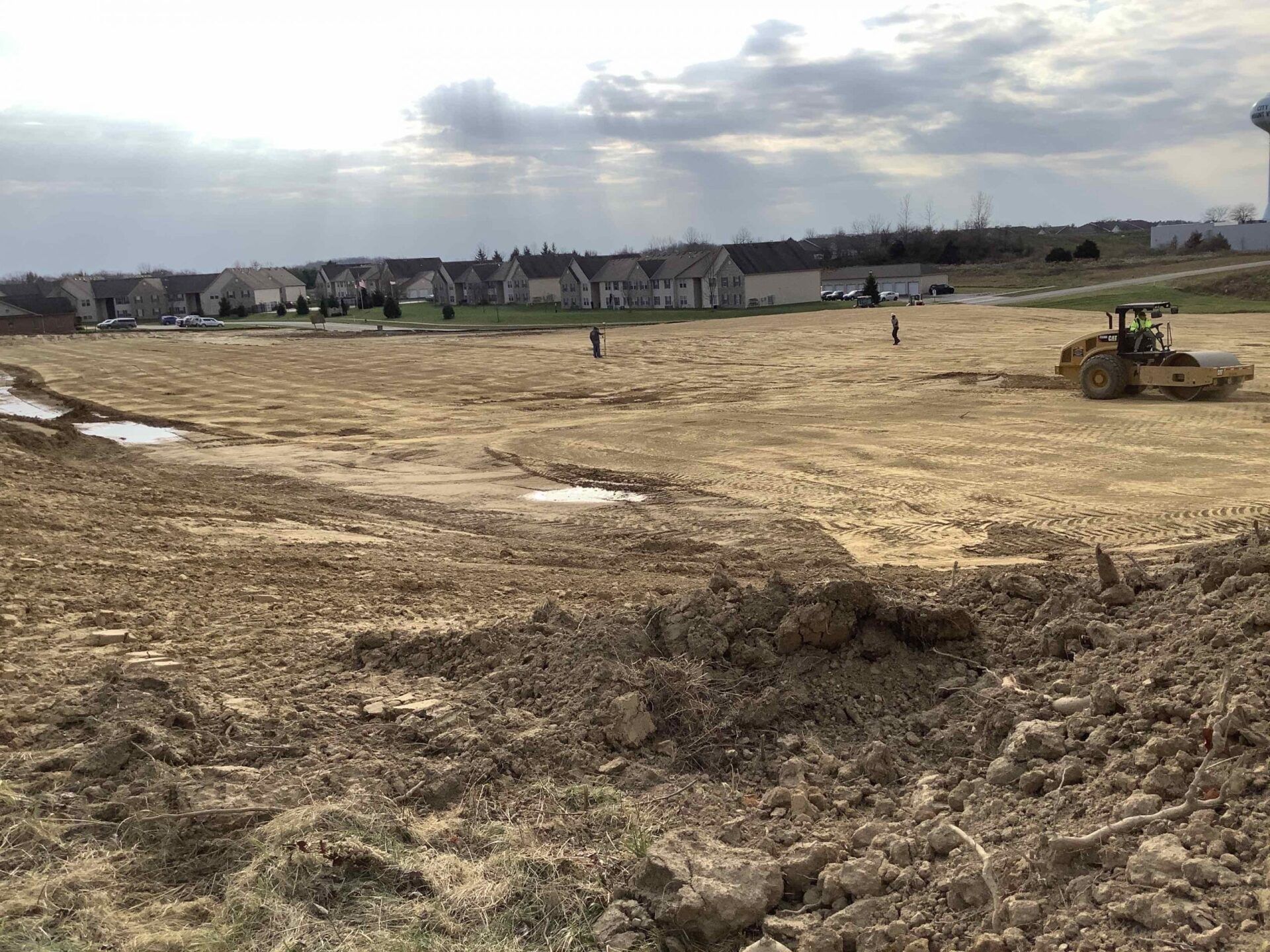 Construction site with heavy equipment, muddy ground, houses in the background under cloudy sky.