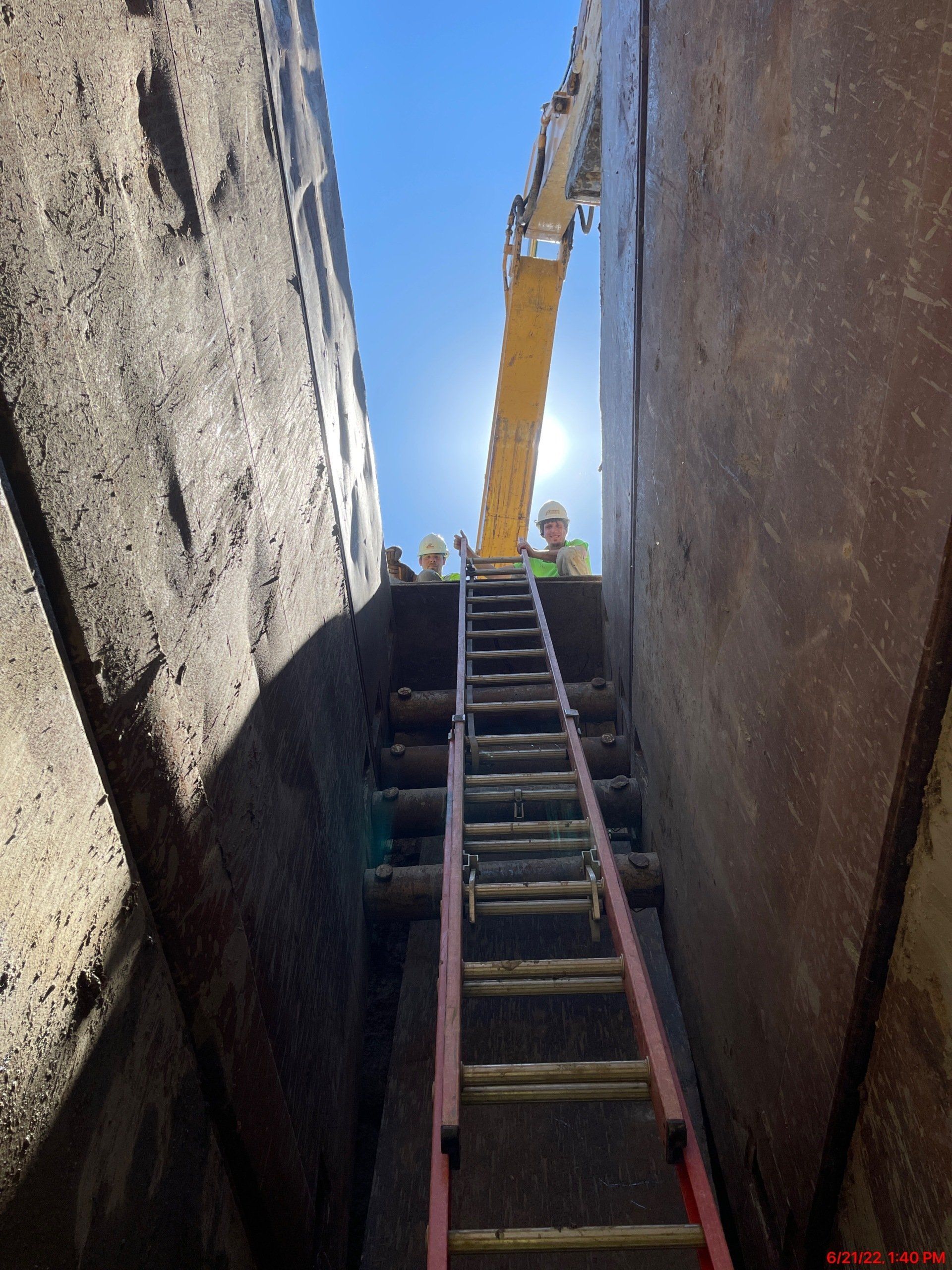 Ladder in deep trench with workers, yellow machinery, and blue sky overhead.