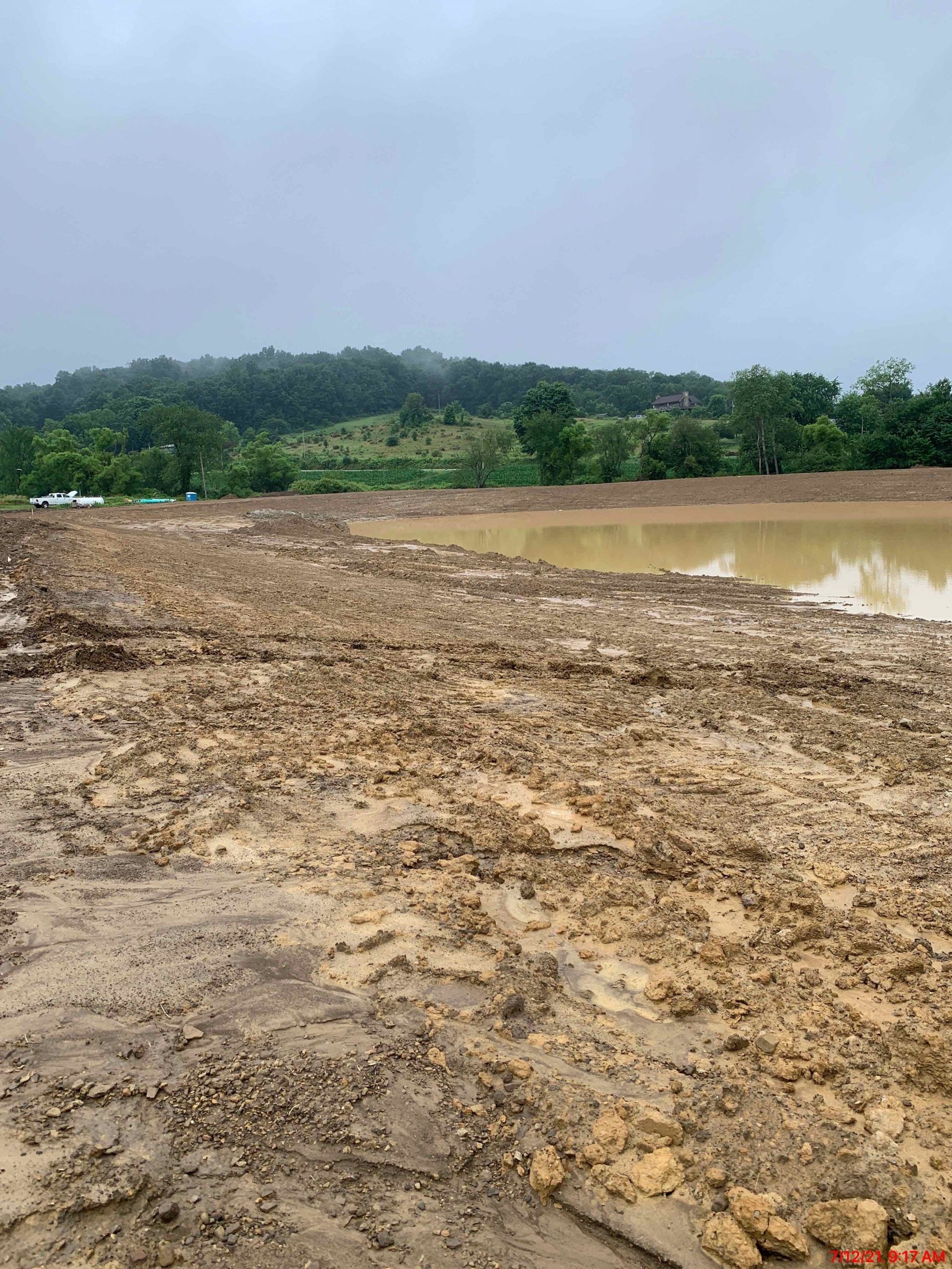 Muddy construction site with a large pond, overcast day, trees in background.
