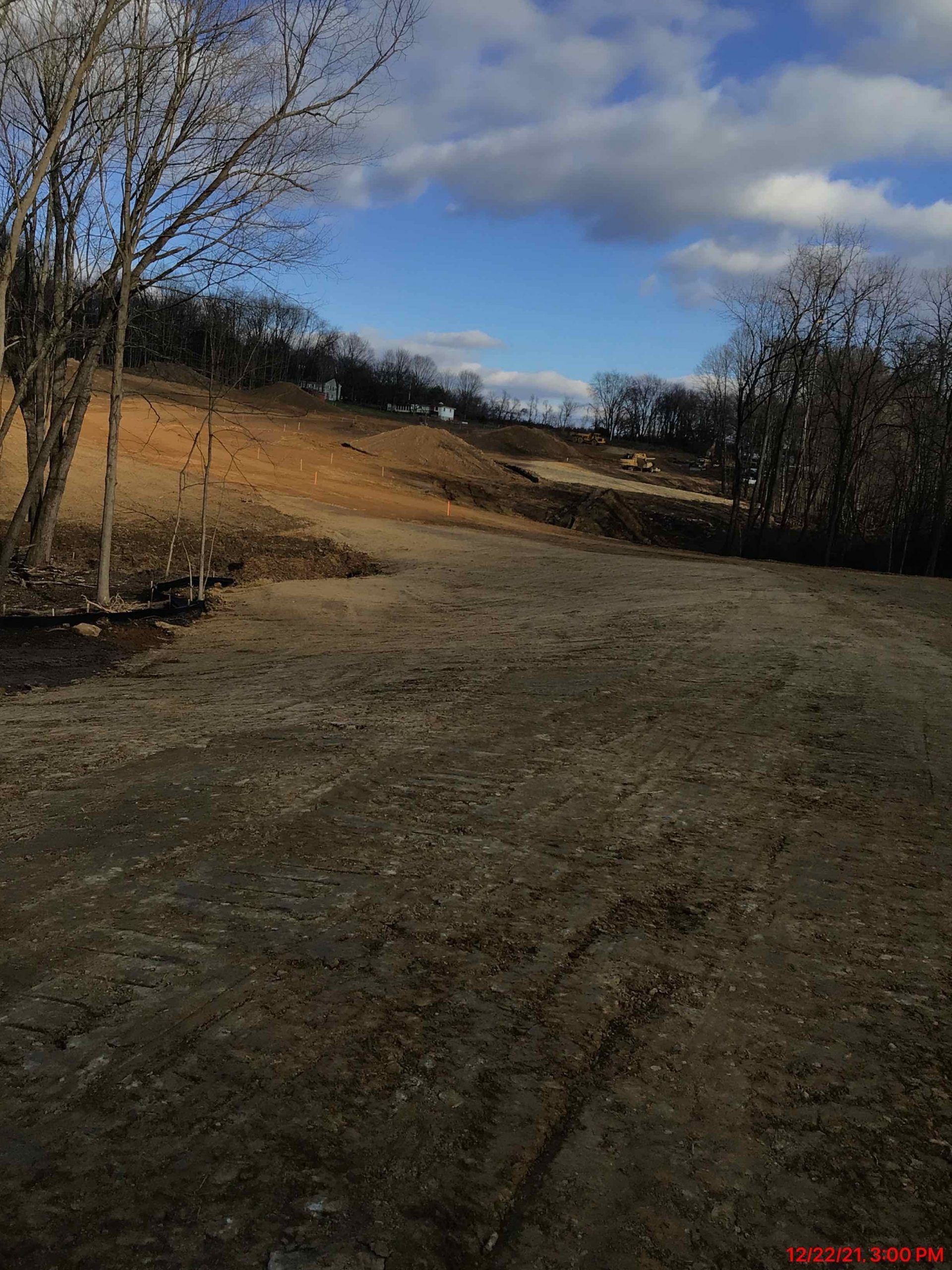 Dirt road leading to a brown hillside under a blue sky with scattered clouds, trees on either side.