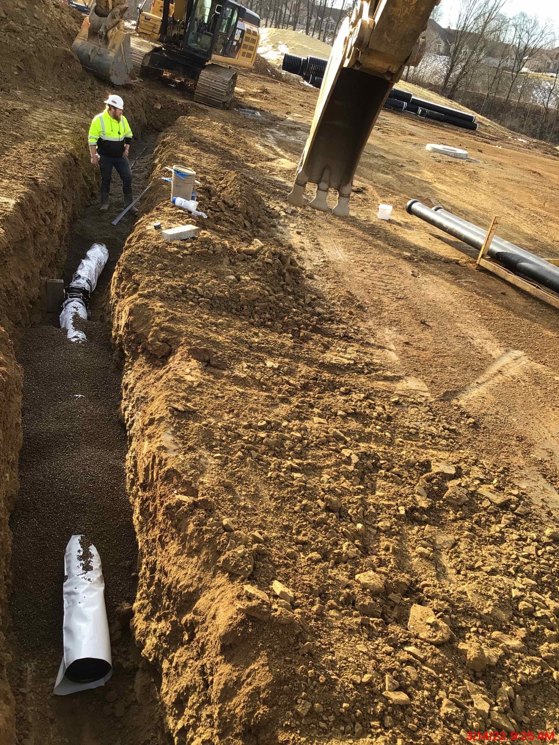 Construction site with a trench, pipes, and worker in safety gear. A backhoe is operating.