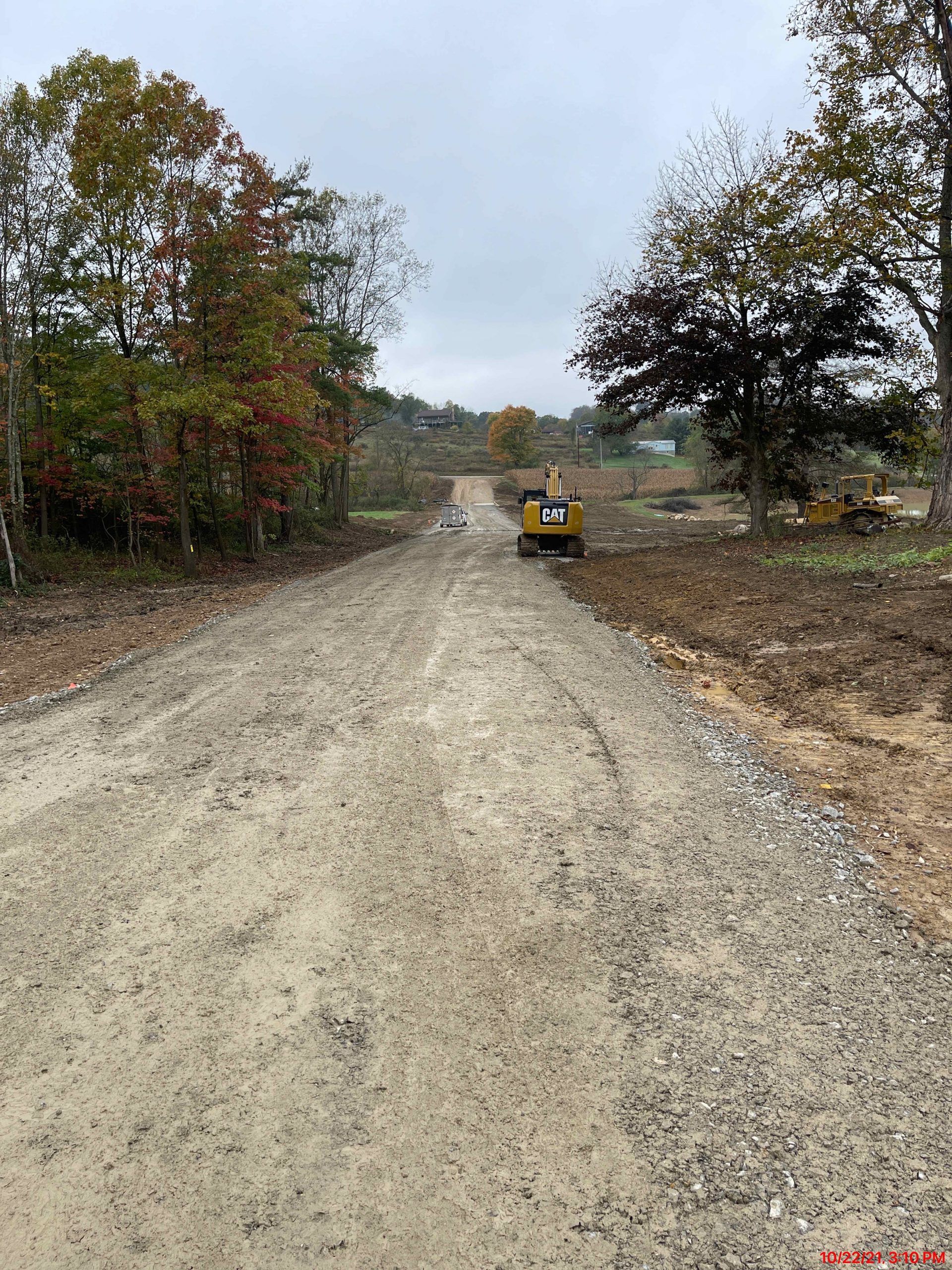 Dirt road leads to construction site with excavator under overcast sky.