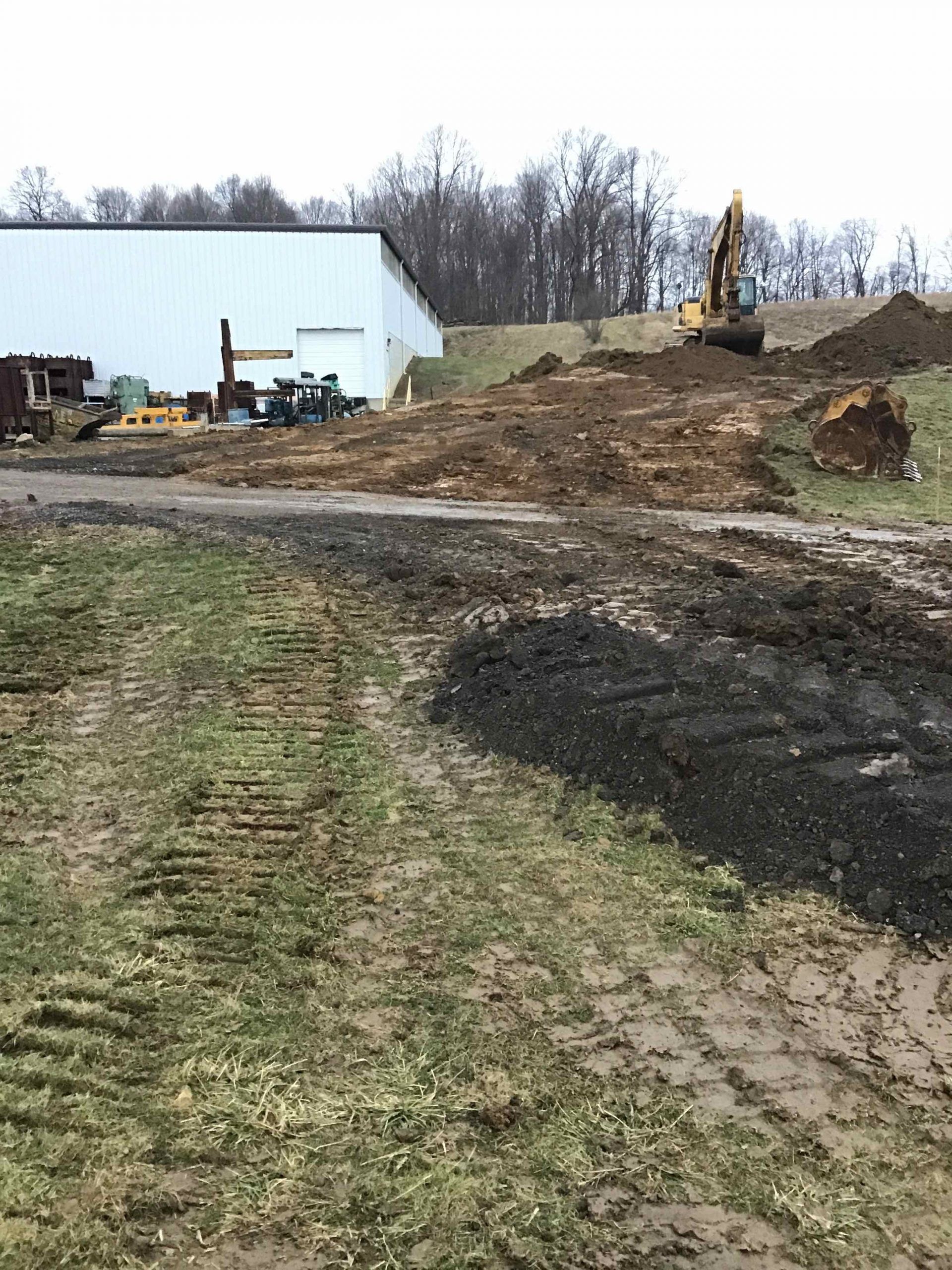 Construction site with an excavator moving dirt near a white building. Grassy foreground, muddy path.