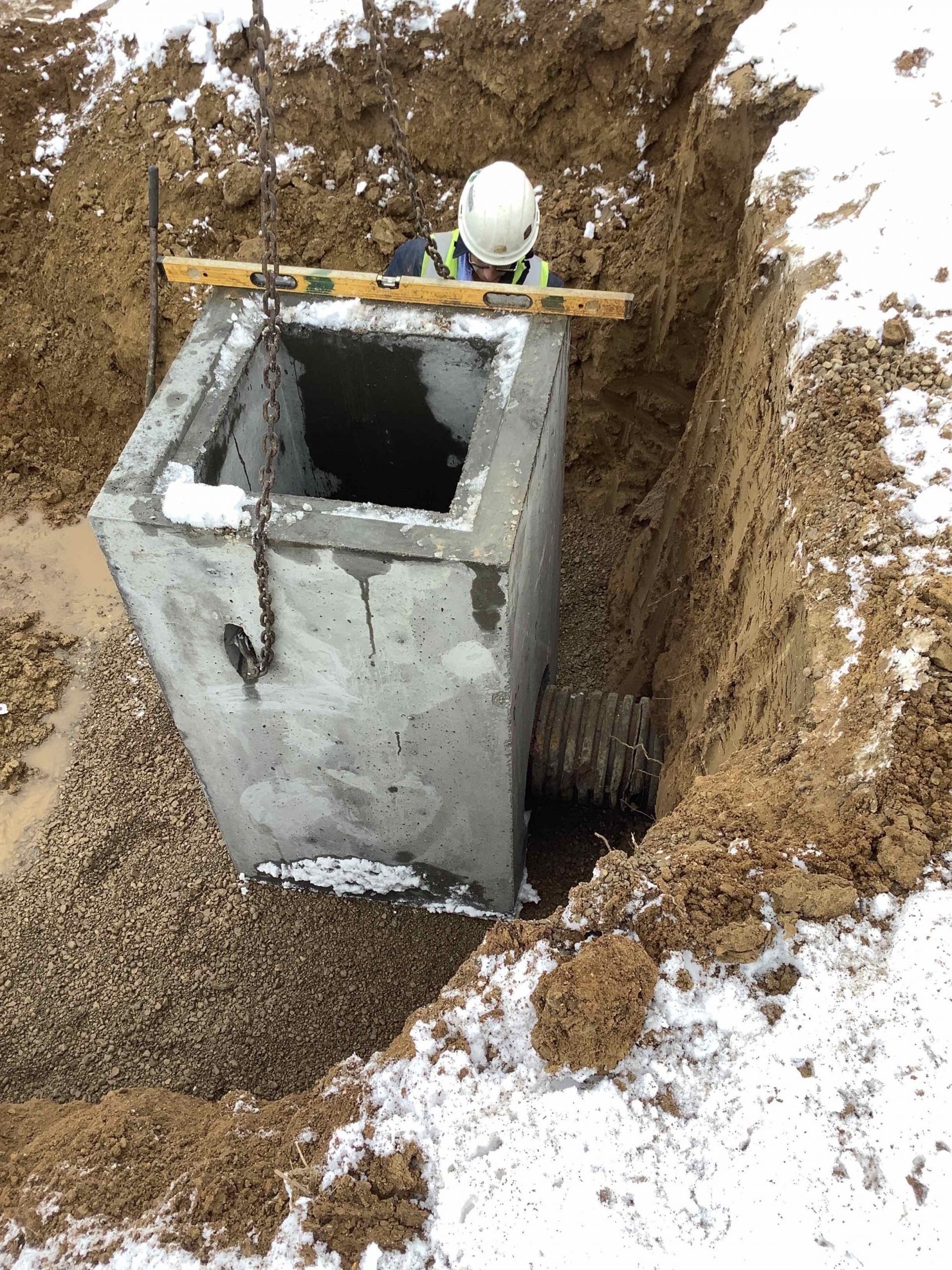 A worker in a trench guides a concrete structure with a crane. Snow covers the ground.