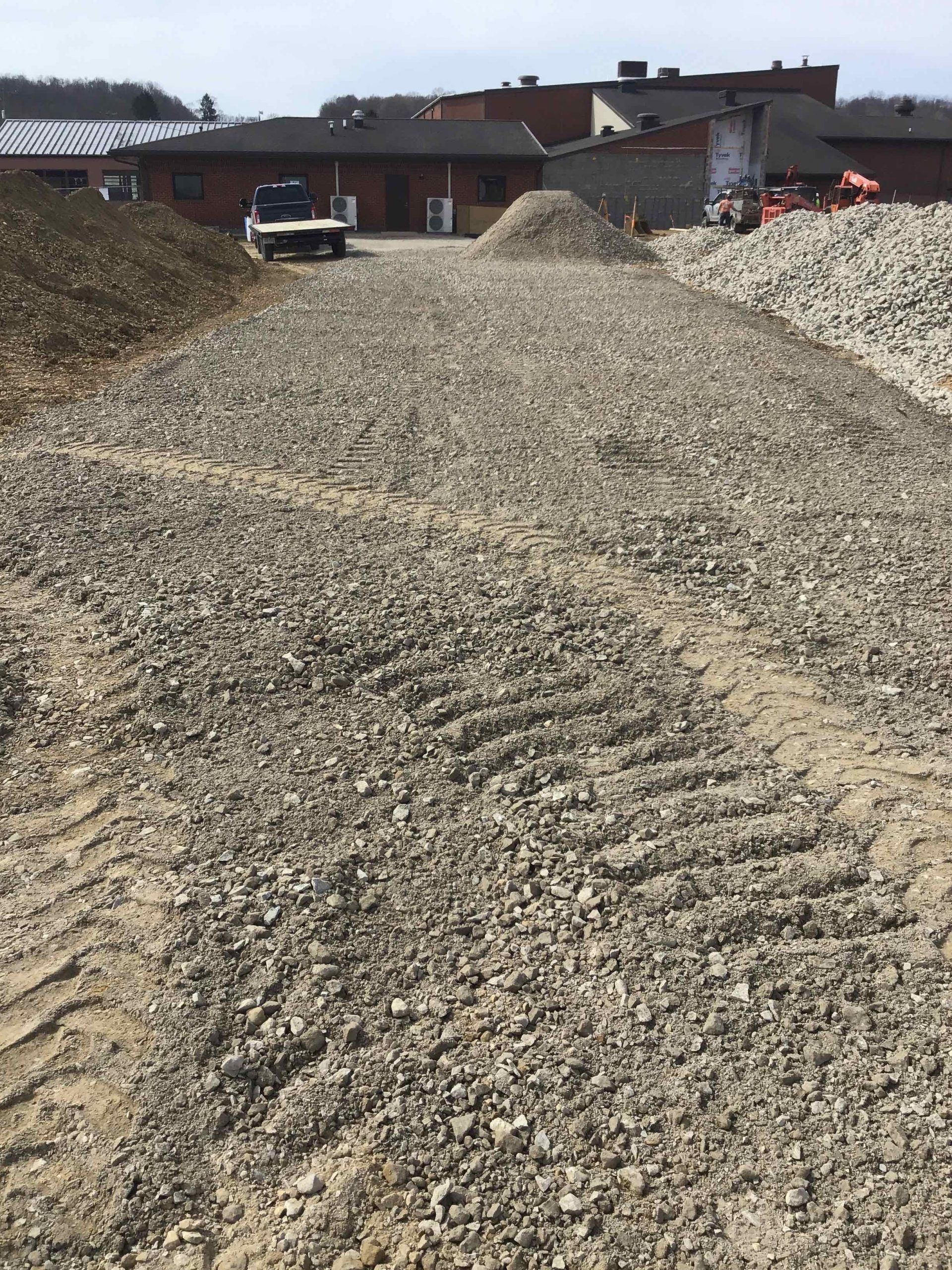 Gravel piles and a path with vehicle tracks. Buildings in the background on a sunny day.