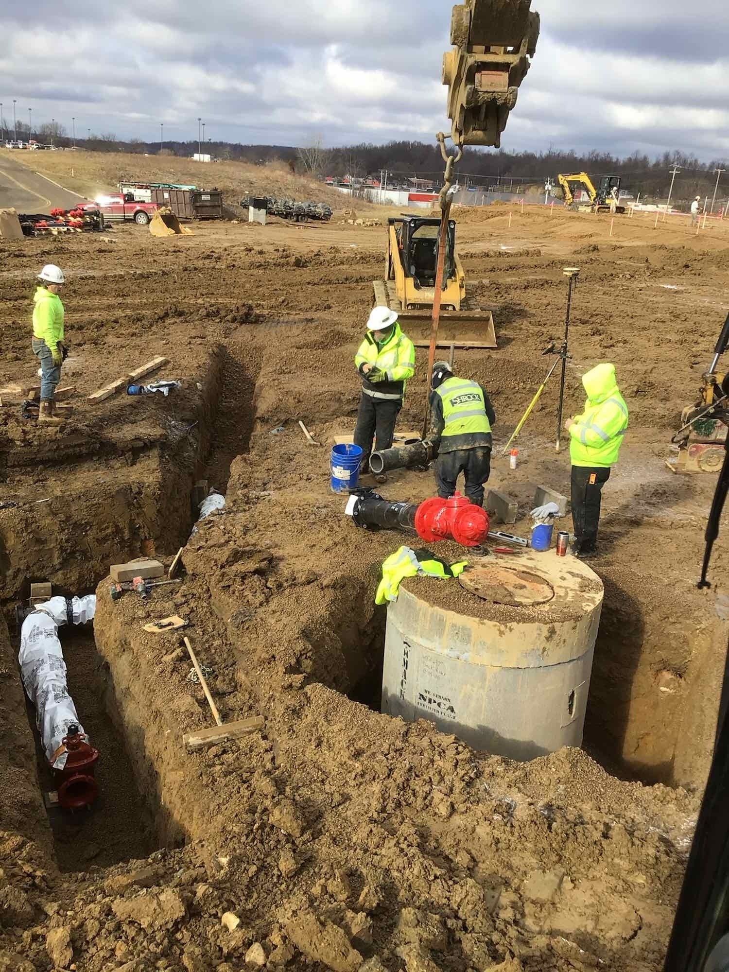 Construction workers in high-vis gear working around open trenches and machinery on a muddy construction site.