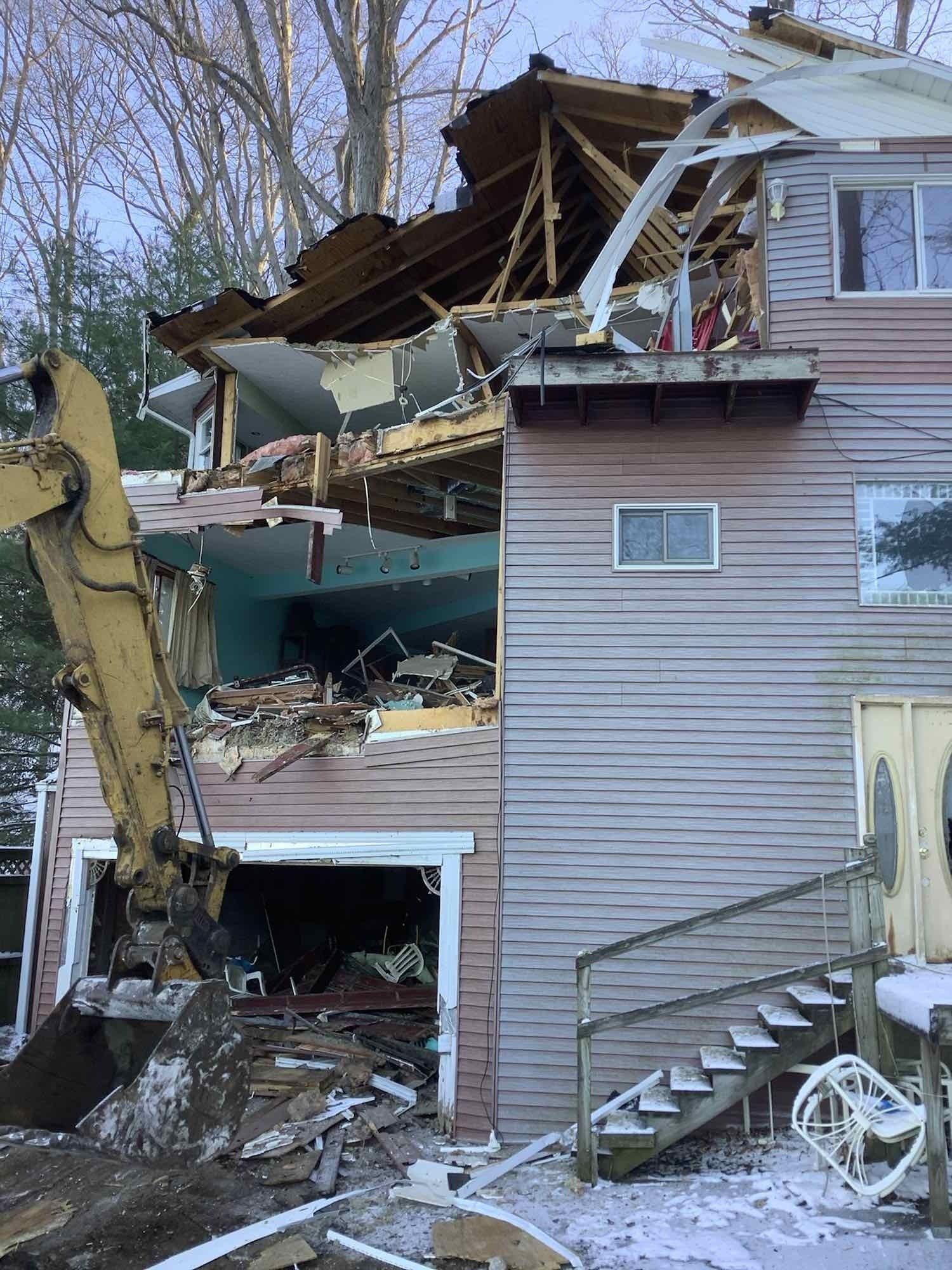A house being demolished by an excavator. Debris and exposed interior visible. Exterior siding is wood.